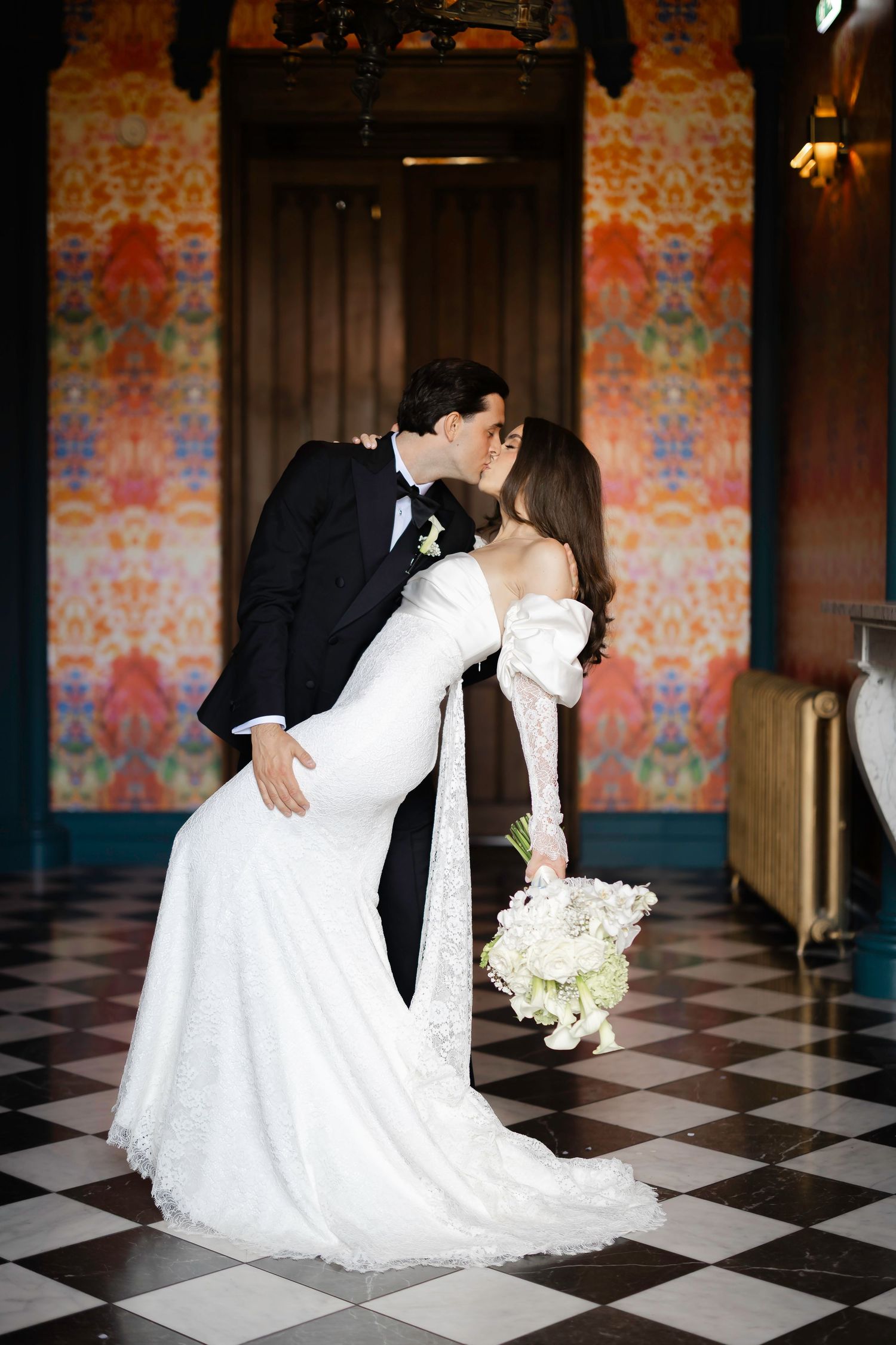 Bride and groom share a romantic kiss in a elegant venue with colorful floral wallpaper and checkered floors.