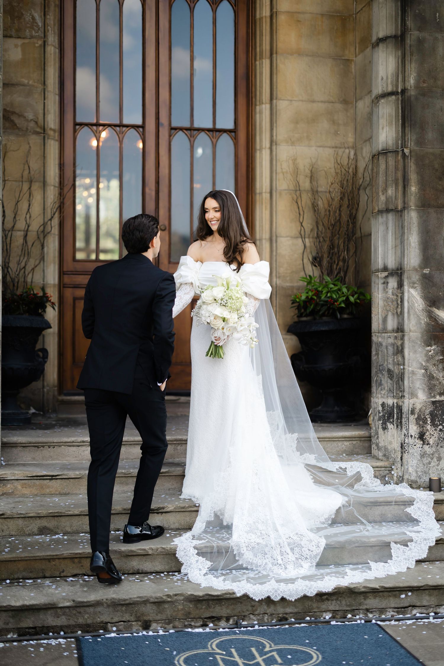 Bride in off-shoulder gown with lace veil holds white bouquet on church steps, facing groom in black tuxedo.