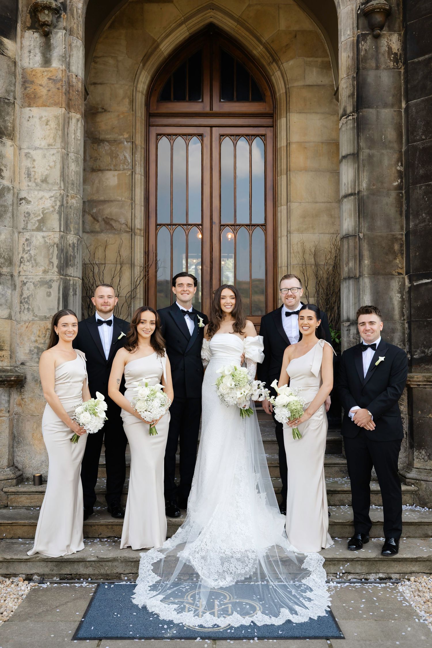 Wedding party poses before Gothic church doors, bride in lace veil gown with bridesmaids in ivory dresses and groomsmen in tuxedos.