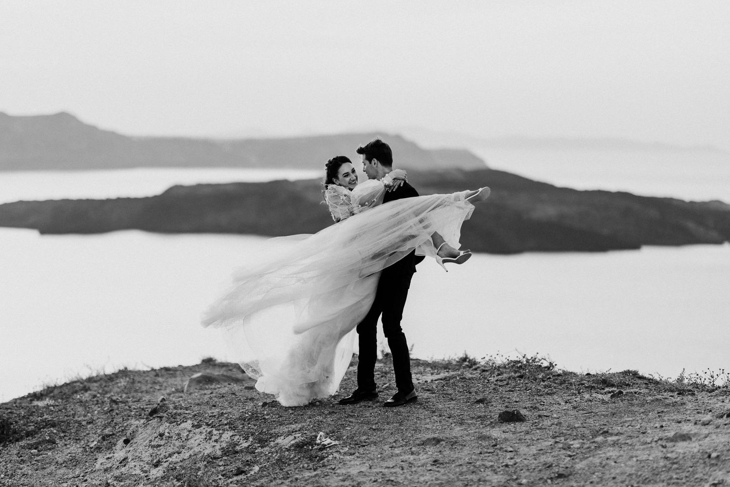 Couple in wedding attire embrace atop a cliff overlooking a serene waterfront vista in black and white.