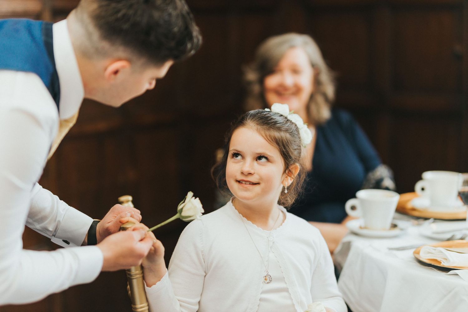 A waiter serves a young guest in a white dress at a restaurant table with coffee cups and plates in the background.