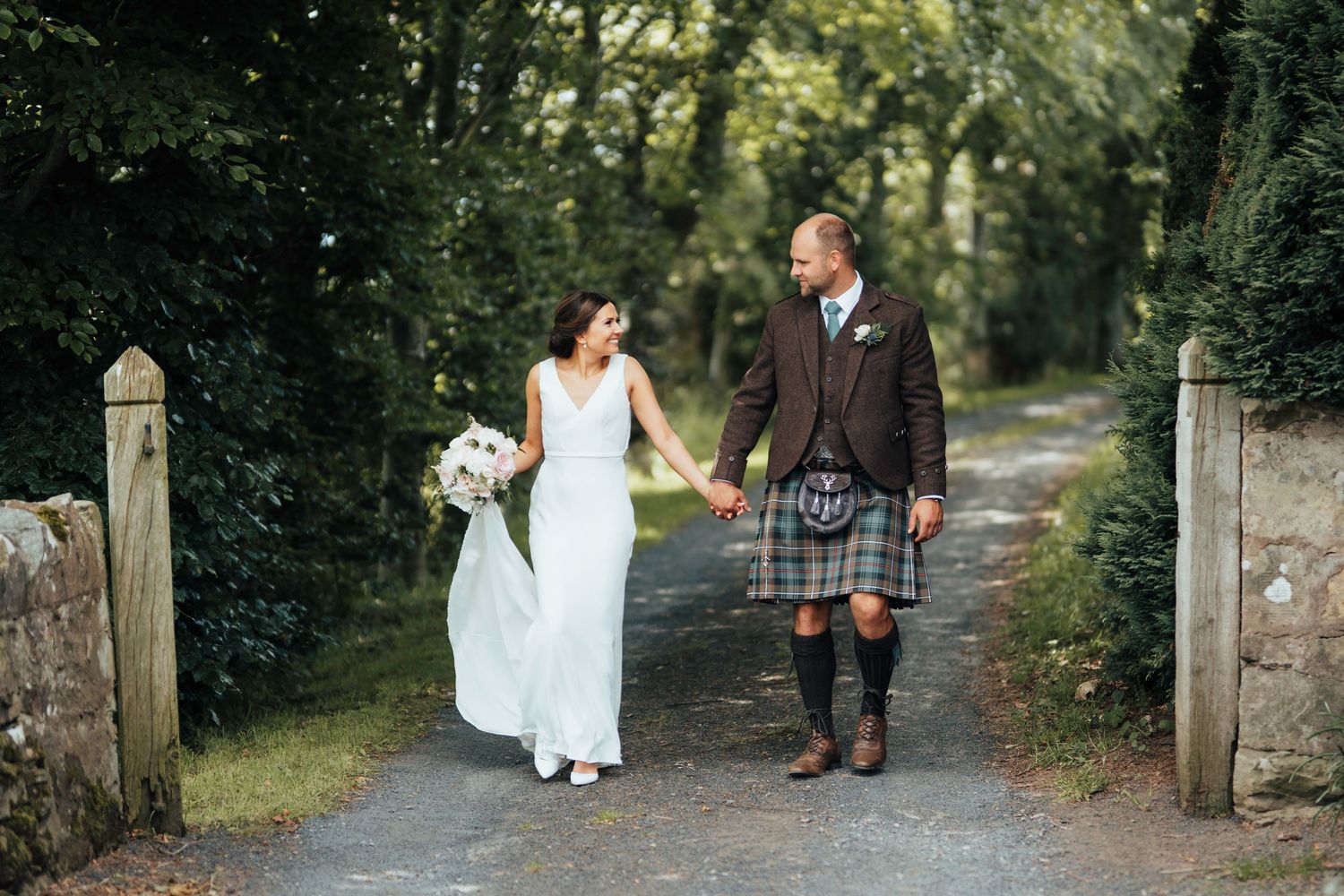 A couple walks together on a woodland path, the groom wearing traditional Scottish kilt and the bride in an elegant white gown.