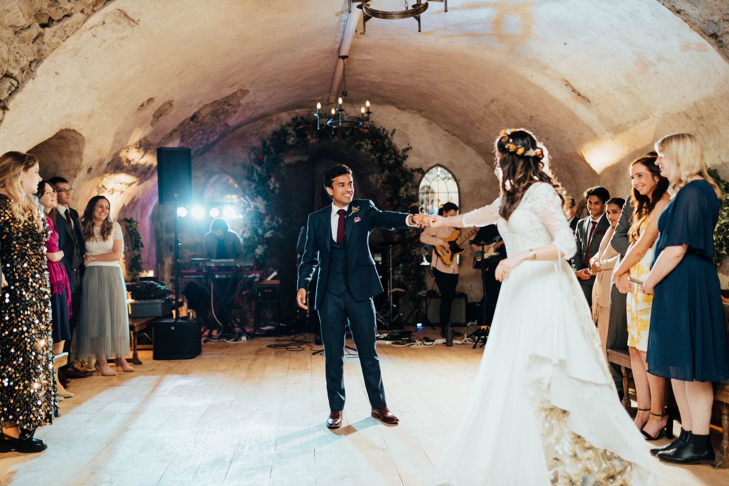 A couple dances on a white floor in a vaulted cellar venue while wedding guests look on.