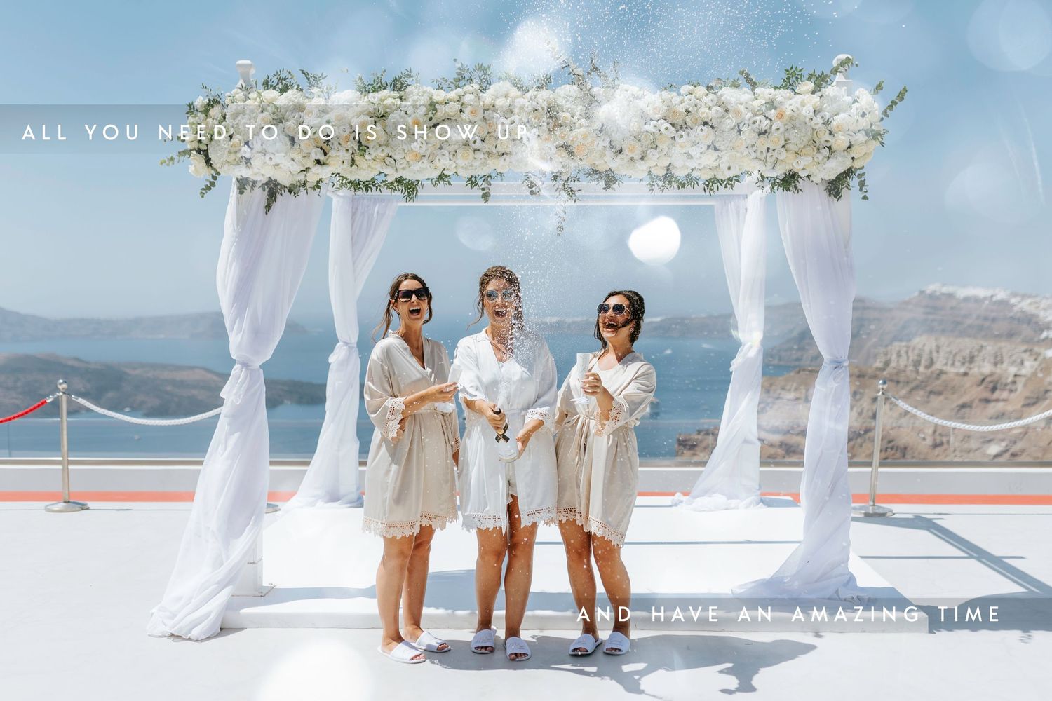 Three people in matching beige outfits pose under a white floral wedding arch with ocean and cliffs in the background.