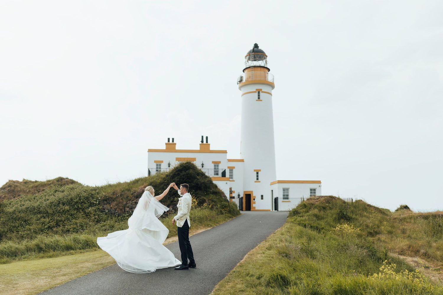 A couple celebrates their wedding day in front of a scenic white lighthouse on a grassy coastal path.