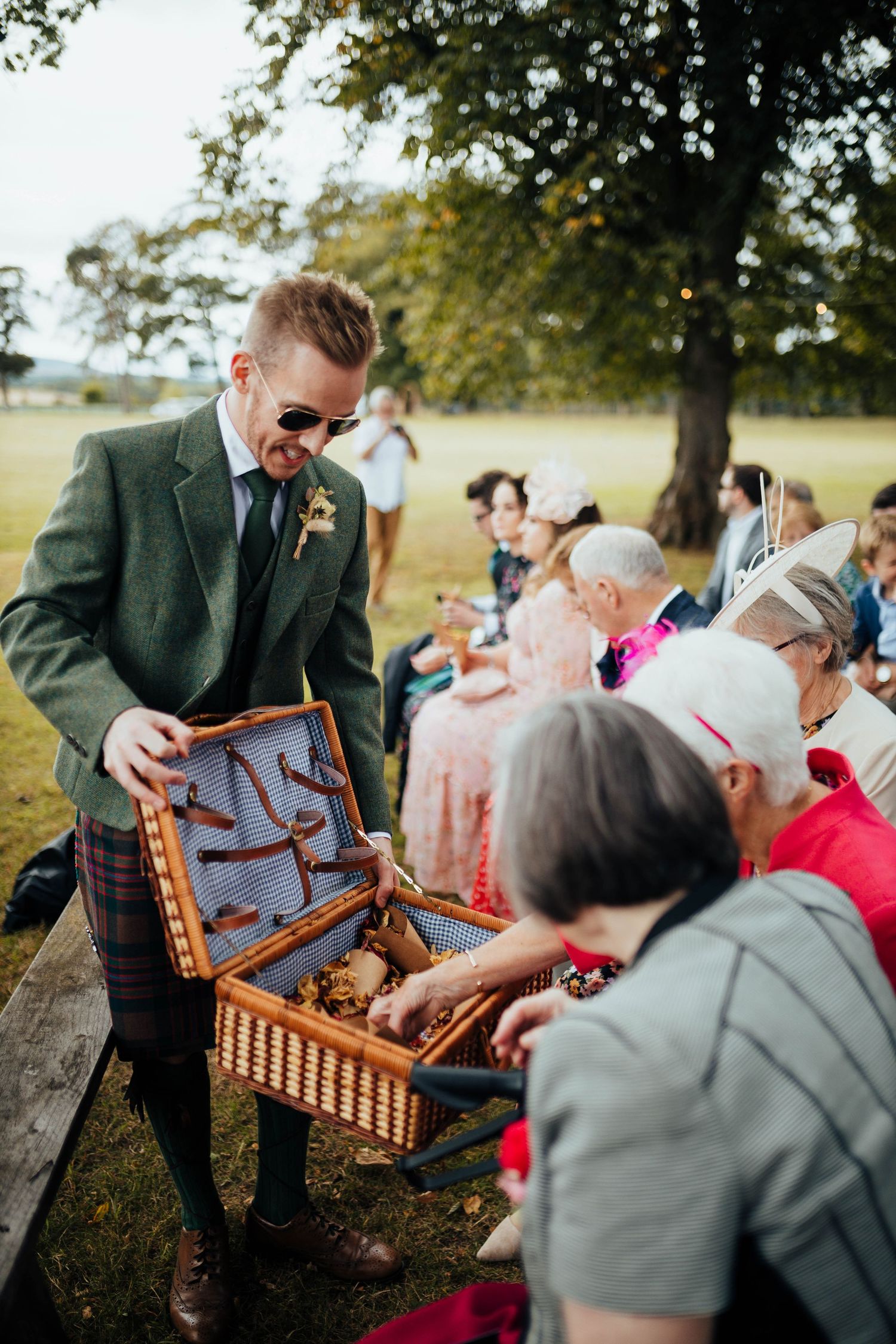 A well-dressed person in a green suit shows a vintage wicker picnic basket to seated guests at an outdoor gathering.