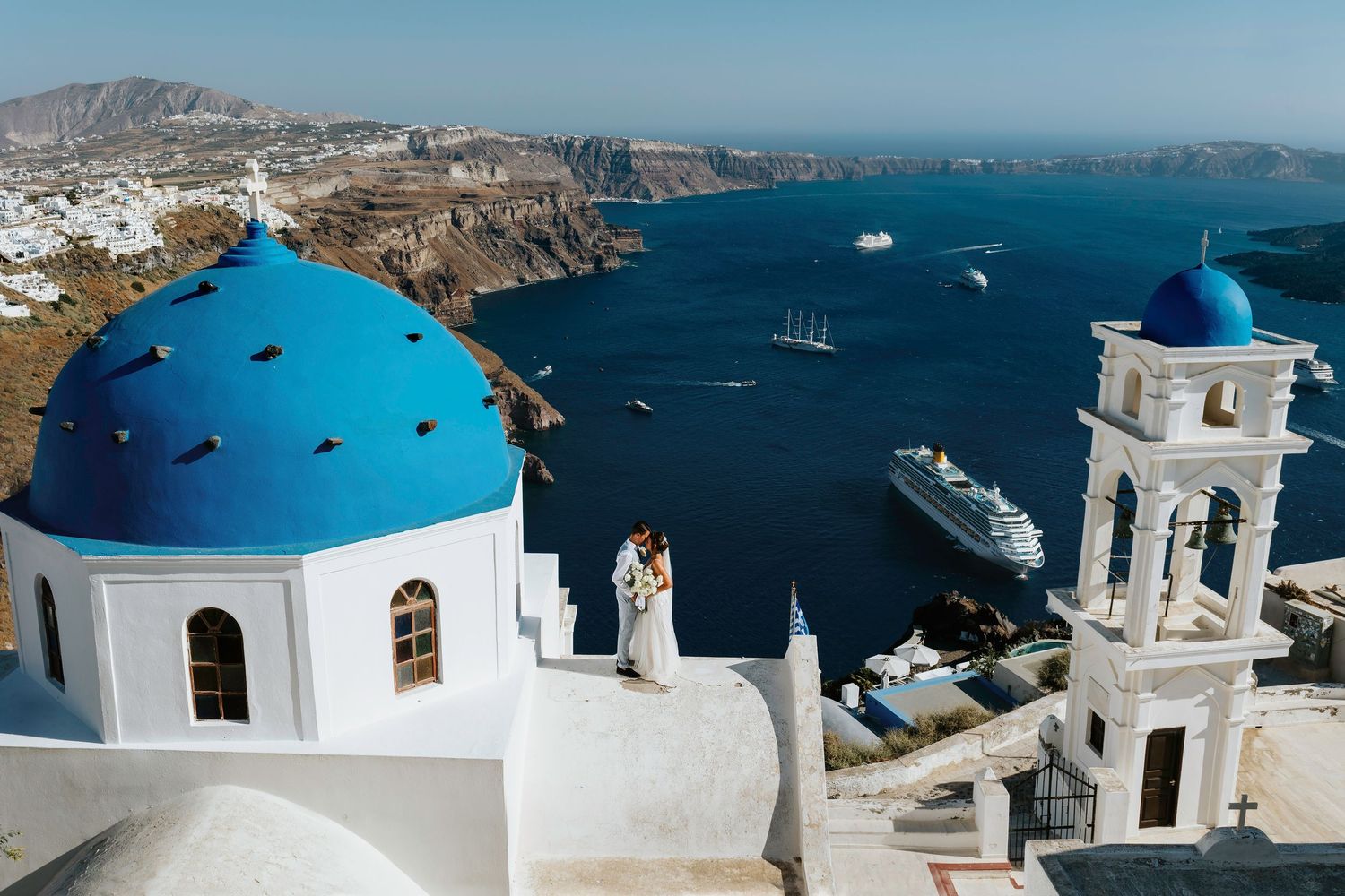 White church with iconic blue dome and bell tower overlooking Santorini's caldera with cruise ships in the Aegean Sea.