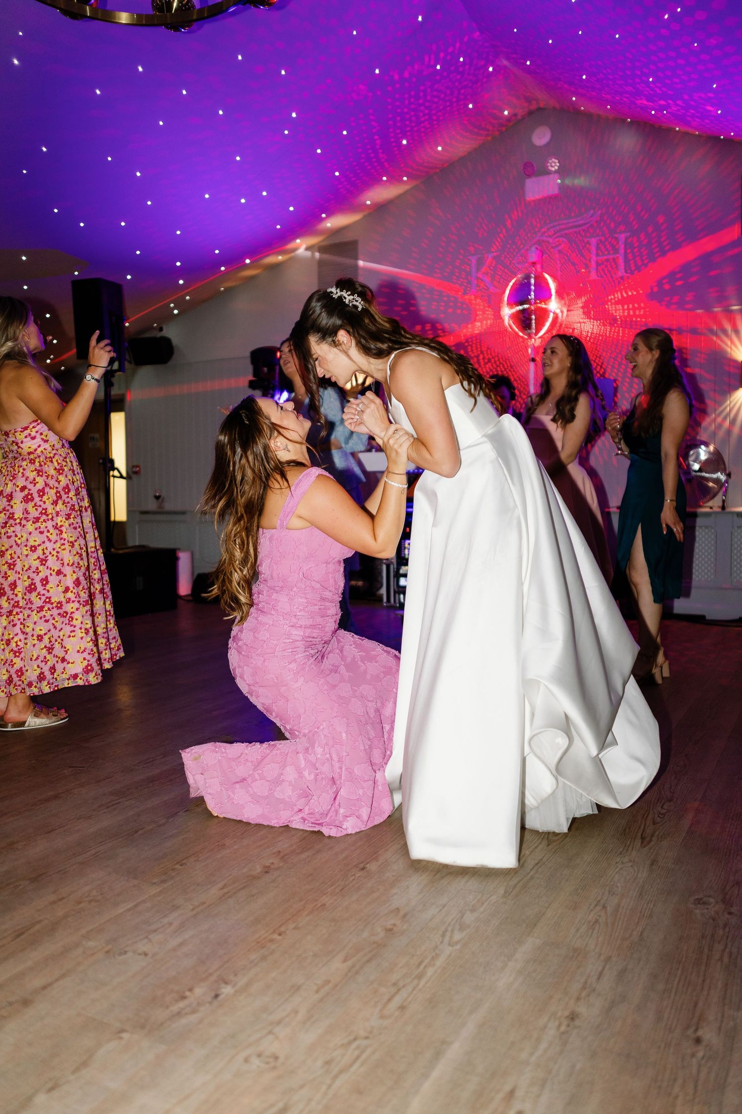 Wedding party guests dance on wooden floor under purple and pink lighting at reception celebration.