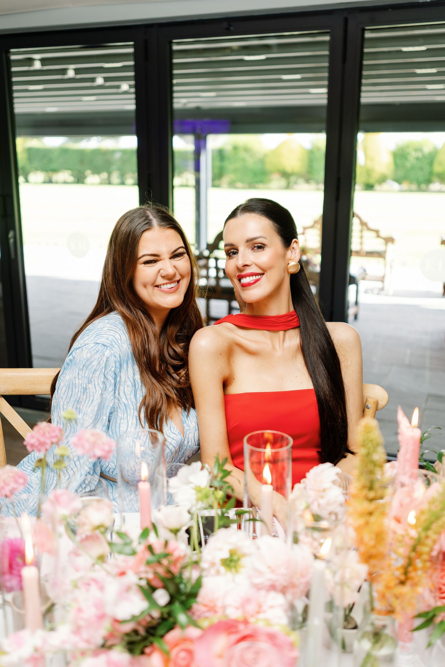 Two friends share a joyful moment at an elegant event with pink floral centerpieces decorating the table.