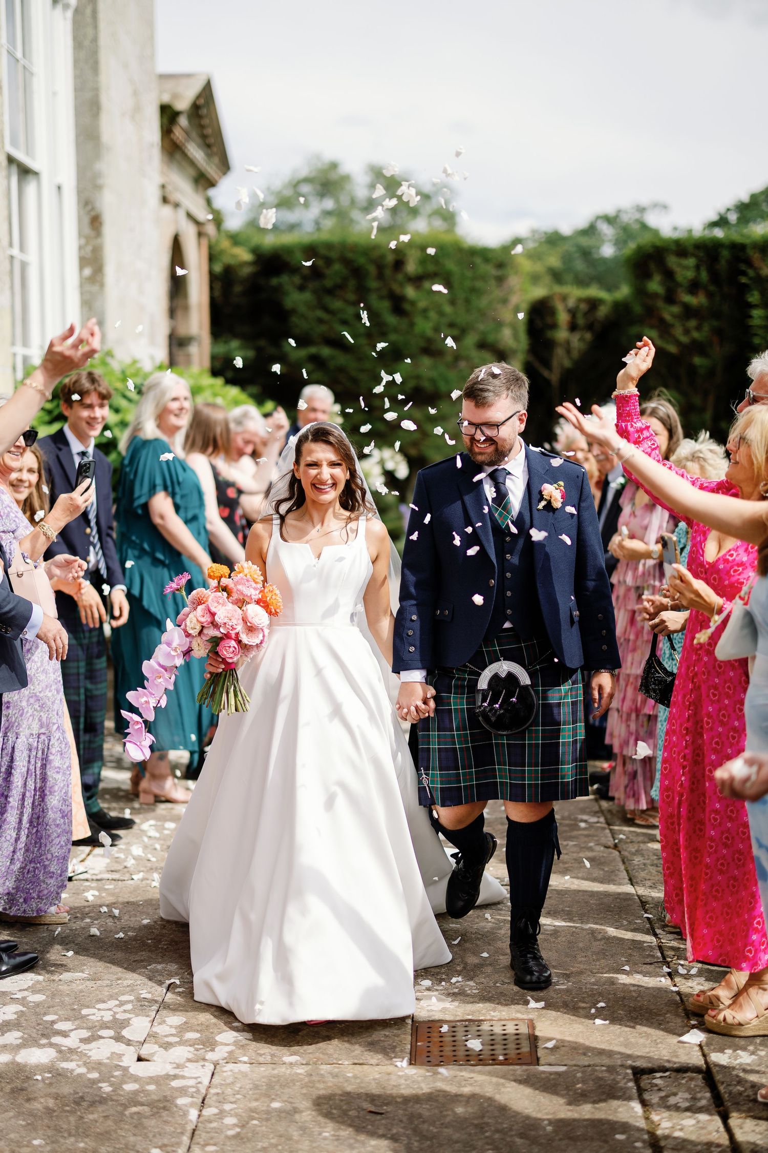Wedding guests throw confetti and celebrate as newlyweds walk through a joyful receiving line outside.
