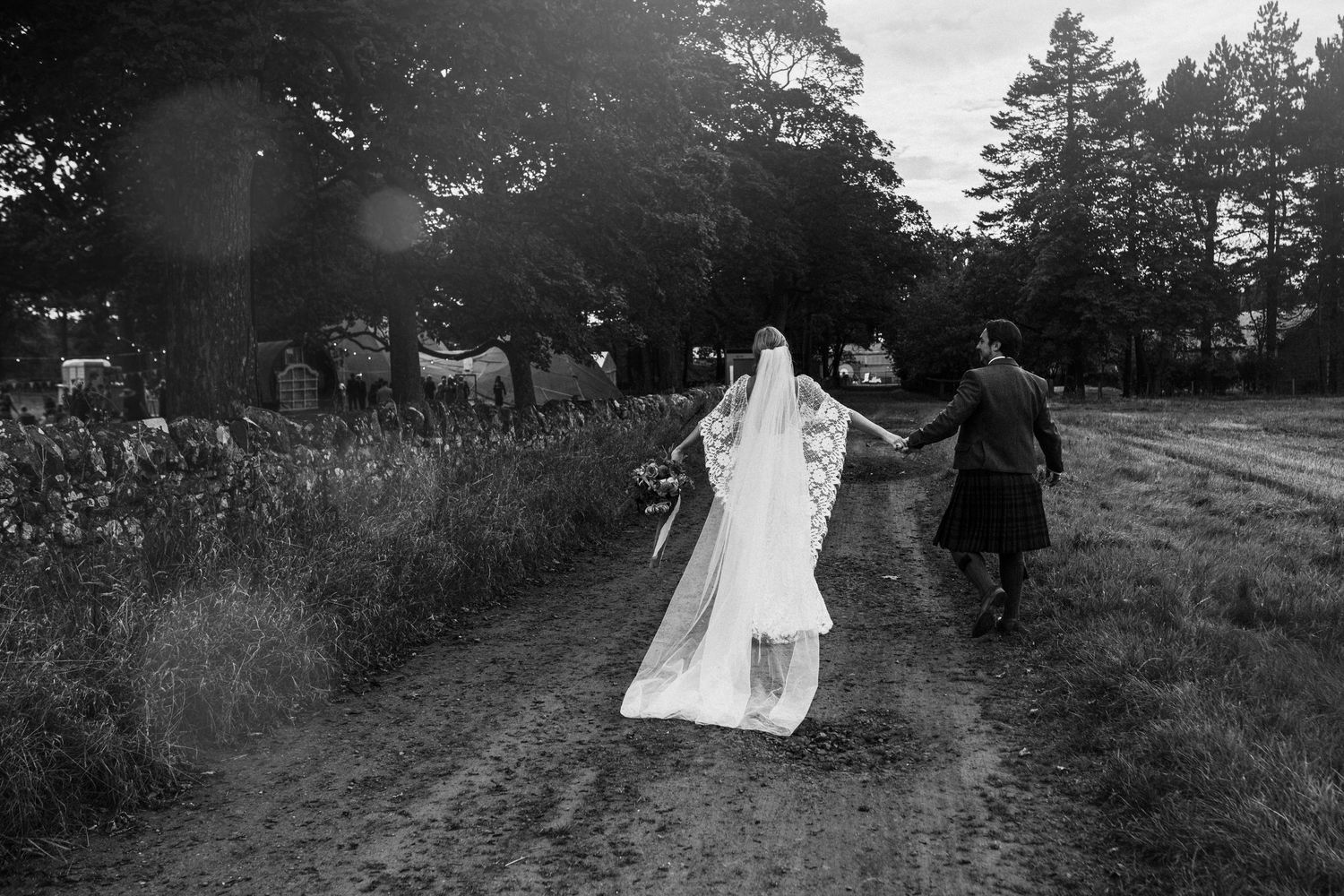 A couple dances on a dirt path surrounded by tall grass and pine trees in a black and white photograph.