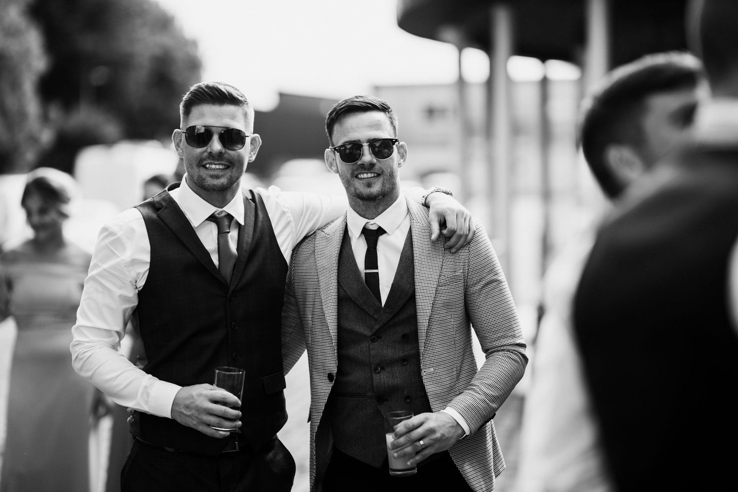 Two men in formal attire wearing waistcoats and sunglasses share drinks at an outdoor event in black and white.