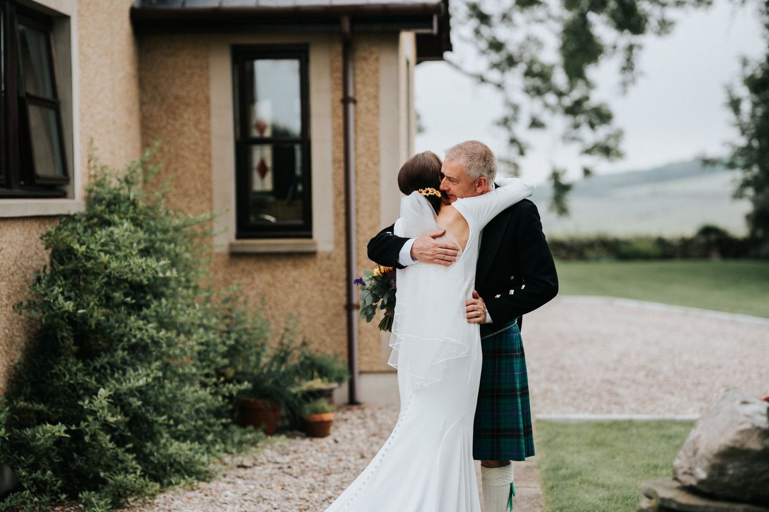 A couple shares a loving embrace during their Scottish wedding outside a stone house in the countryside.