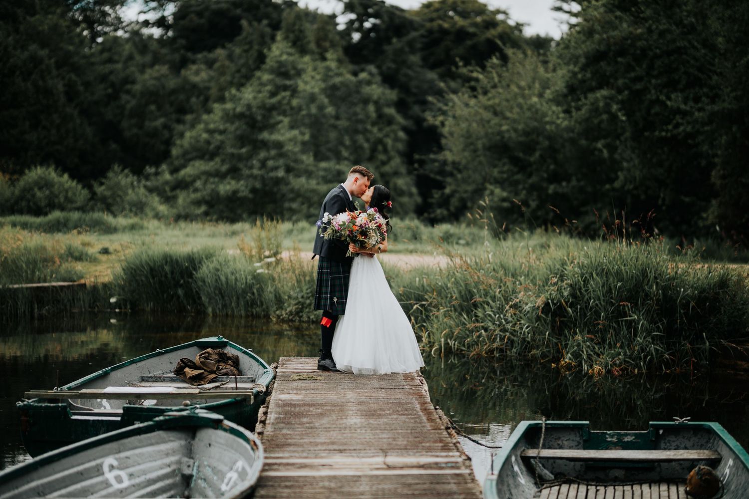 Newlywed couple embraces on wooden dock surrounded by boats and lush greenery in romantic outdoor setting.
