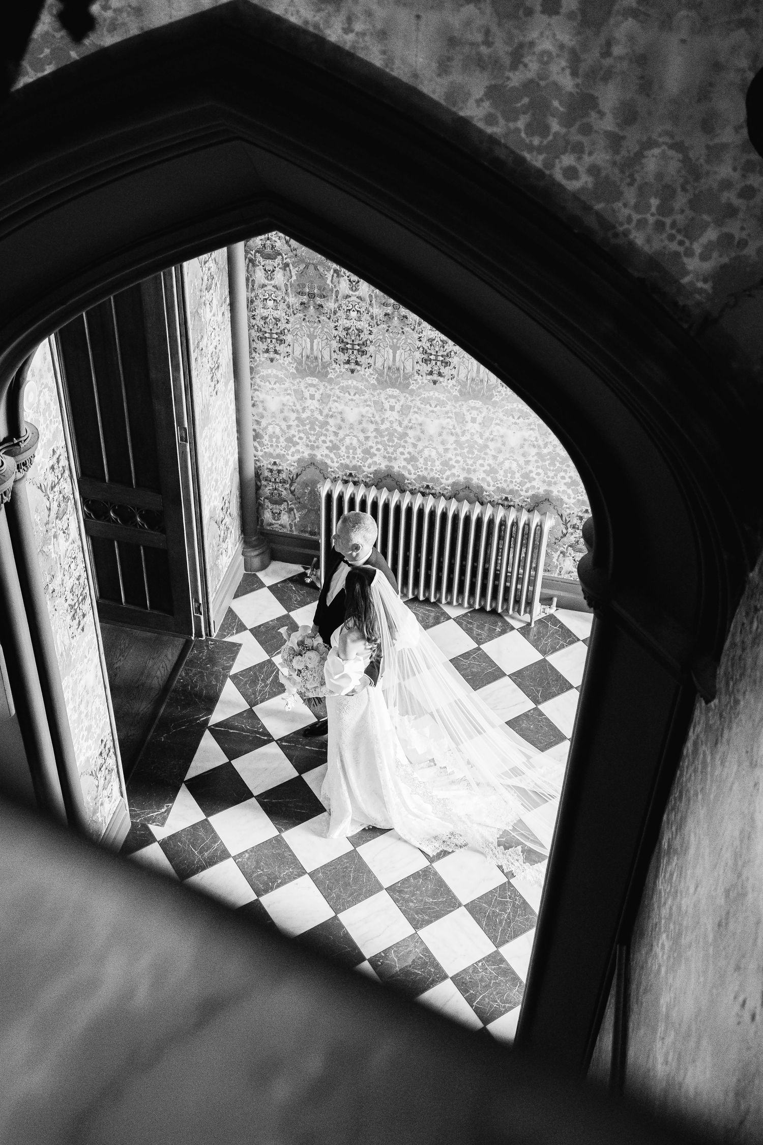 Black and white wedding photo of bride and groom framed by gothic arch on checkered floor.