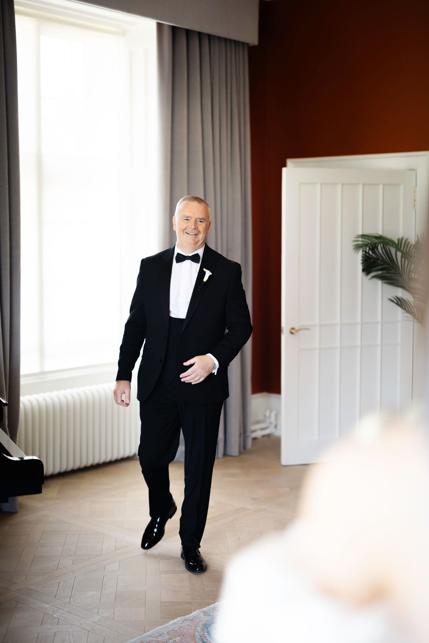 A smiling groom in a black tuxedo with bow tie and boutonniere stands in an elegant room on his wedding day.