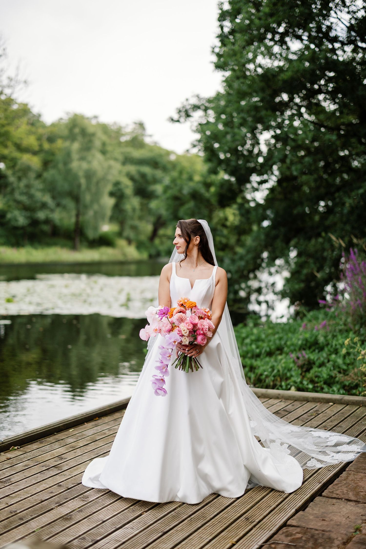 Bride in white wedding dress with colorful bouquet sits gracefully on wooden dock by serene lake surrounded by greenery.