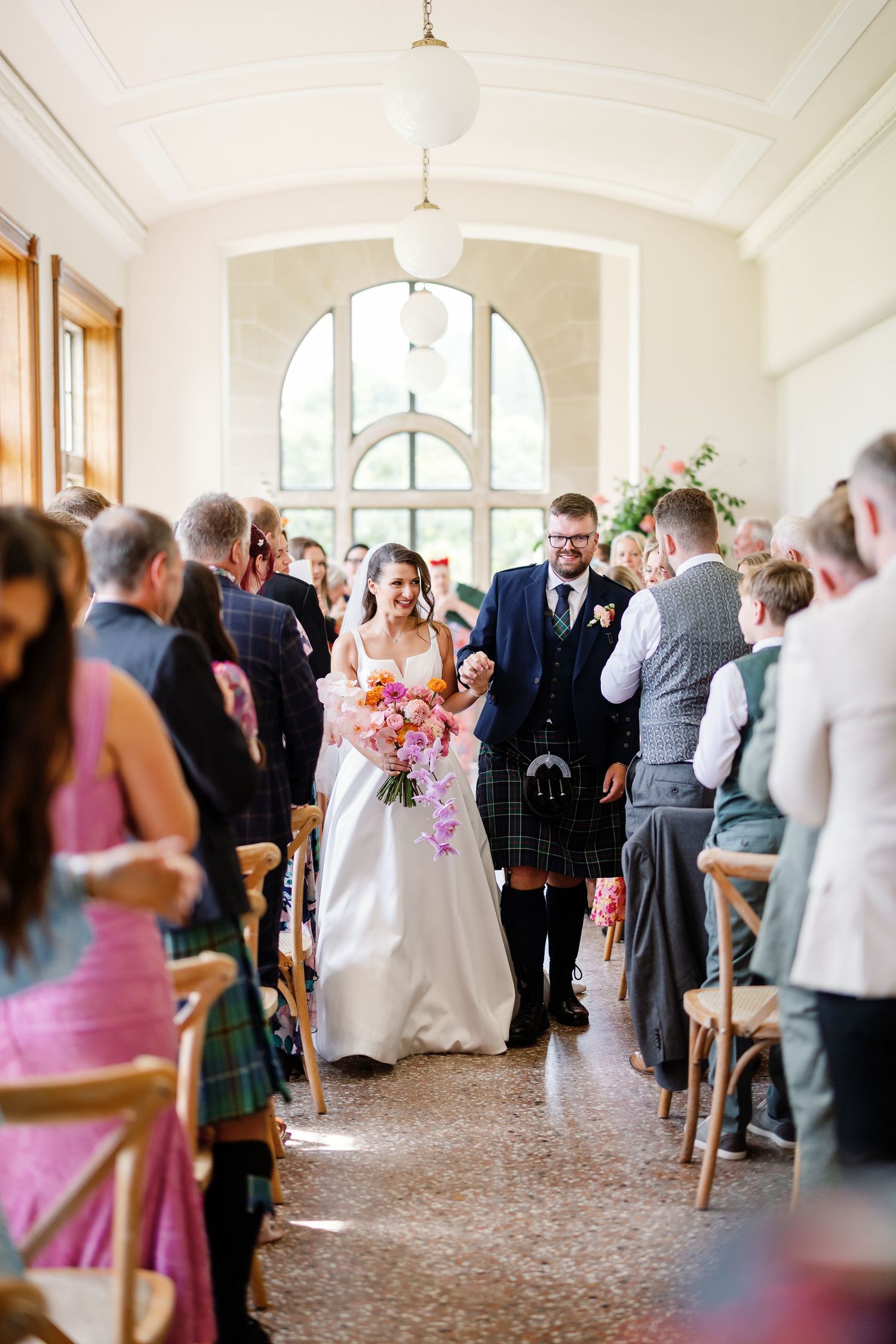 A wedding ceremony in a bright indoor venue with arched windows and guests seated on wooden chairs.