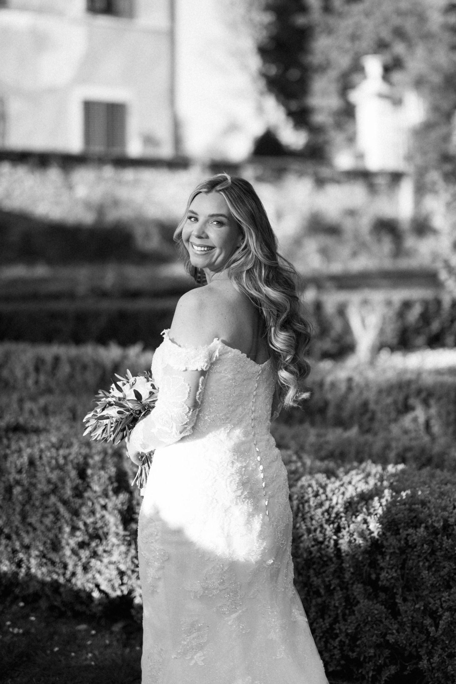 A radiant bride in a lace off-shoulder gown poses in a garden setting in black and white photography villa catignano wedding pictures