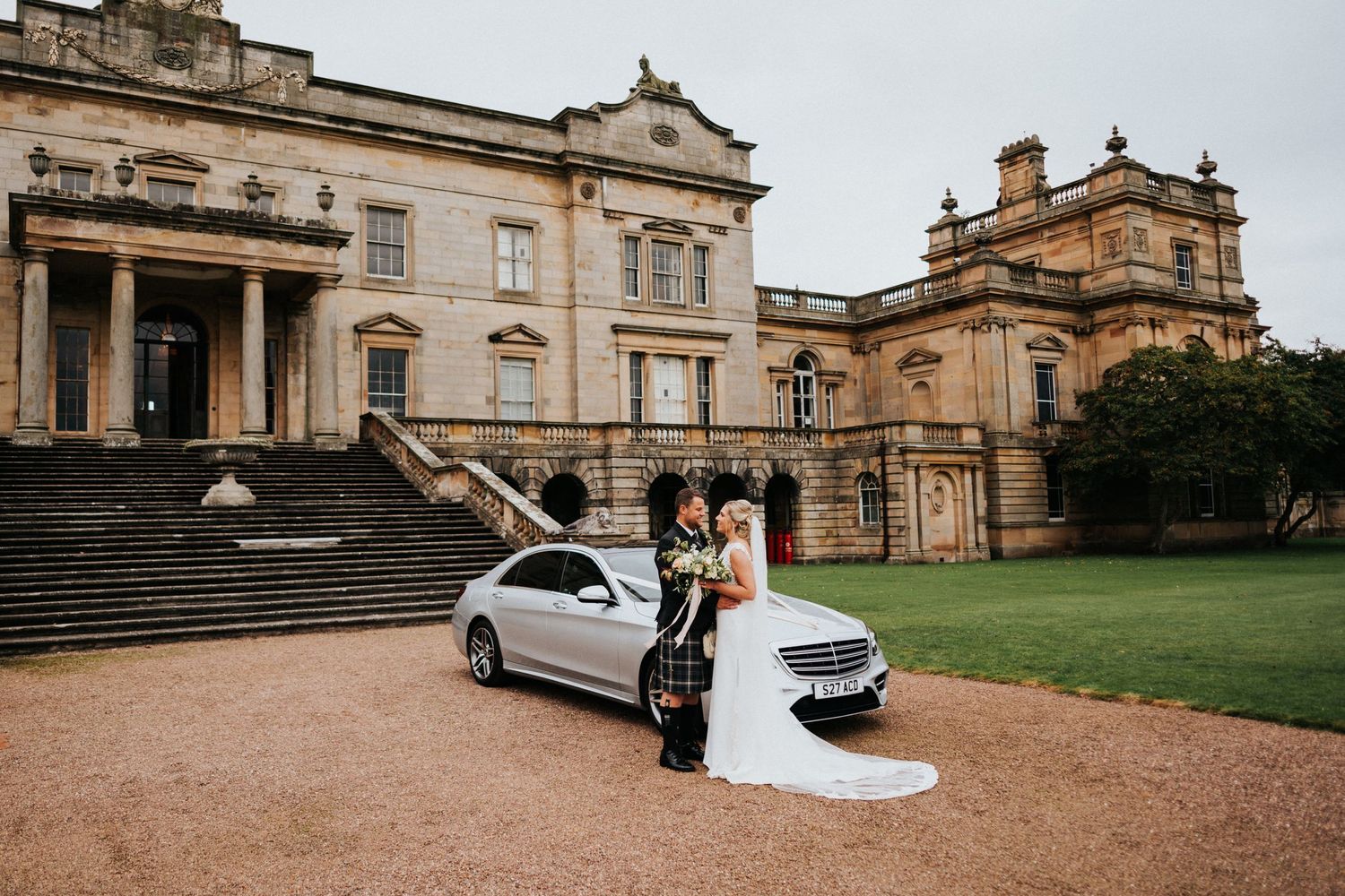 Newlyweds celebrate next to a silver car outside an elegant historic mansion with grand stone steps and architecture.