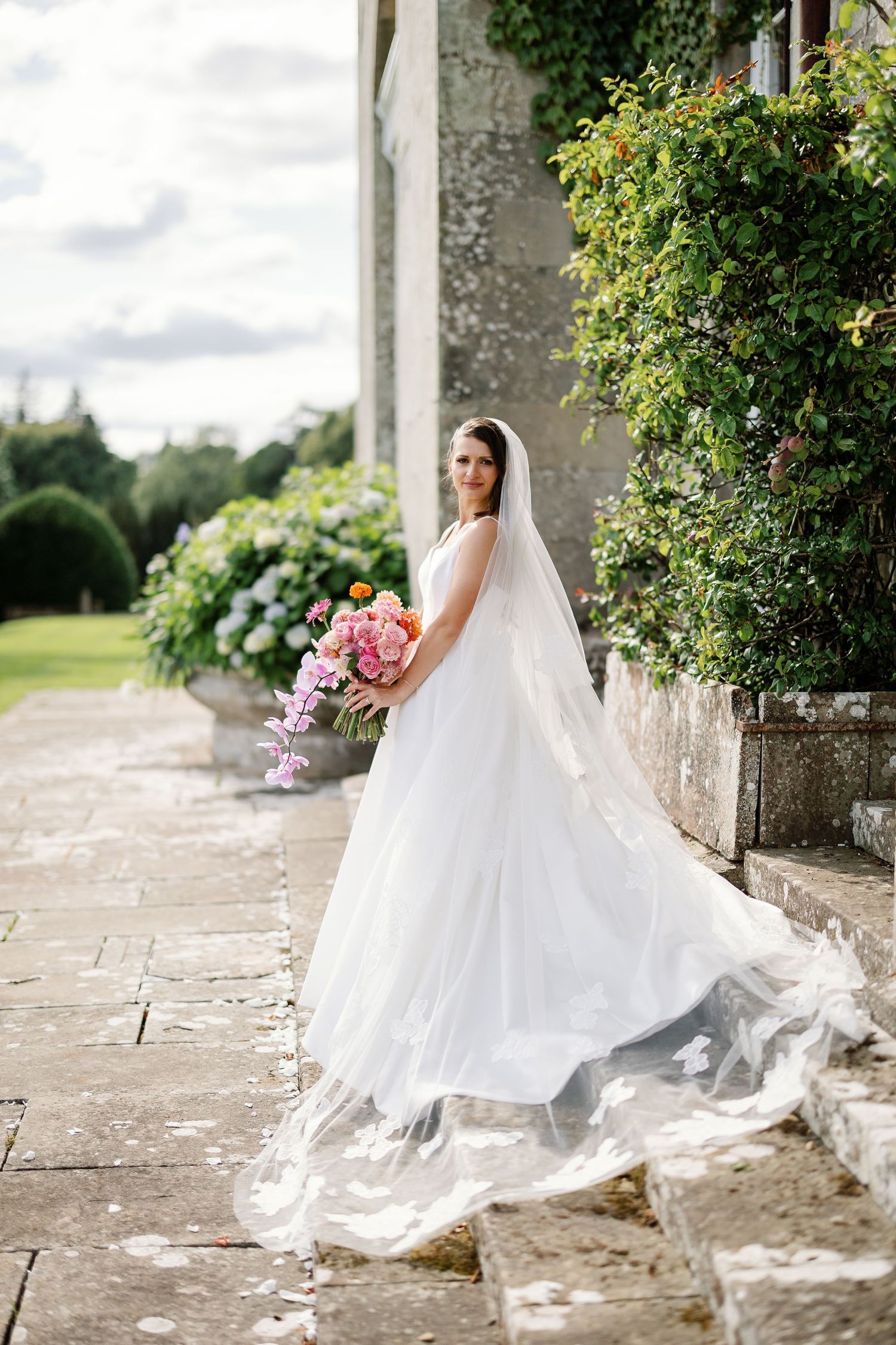 A bride in a flowing white wedding dress sits gracefully on stone steps surrounded by lush greenery and flowers.