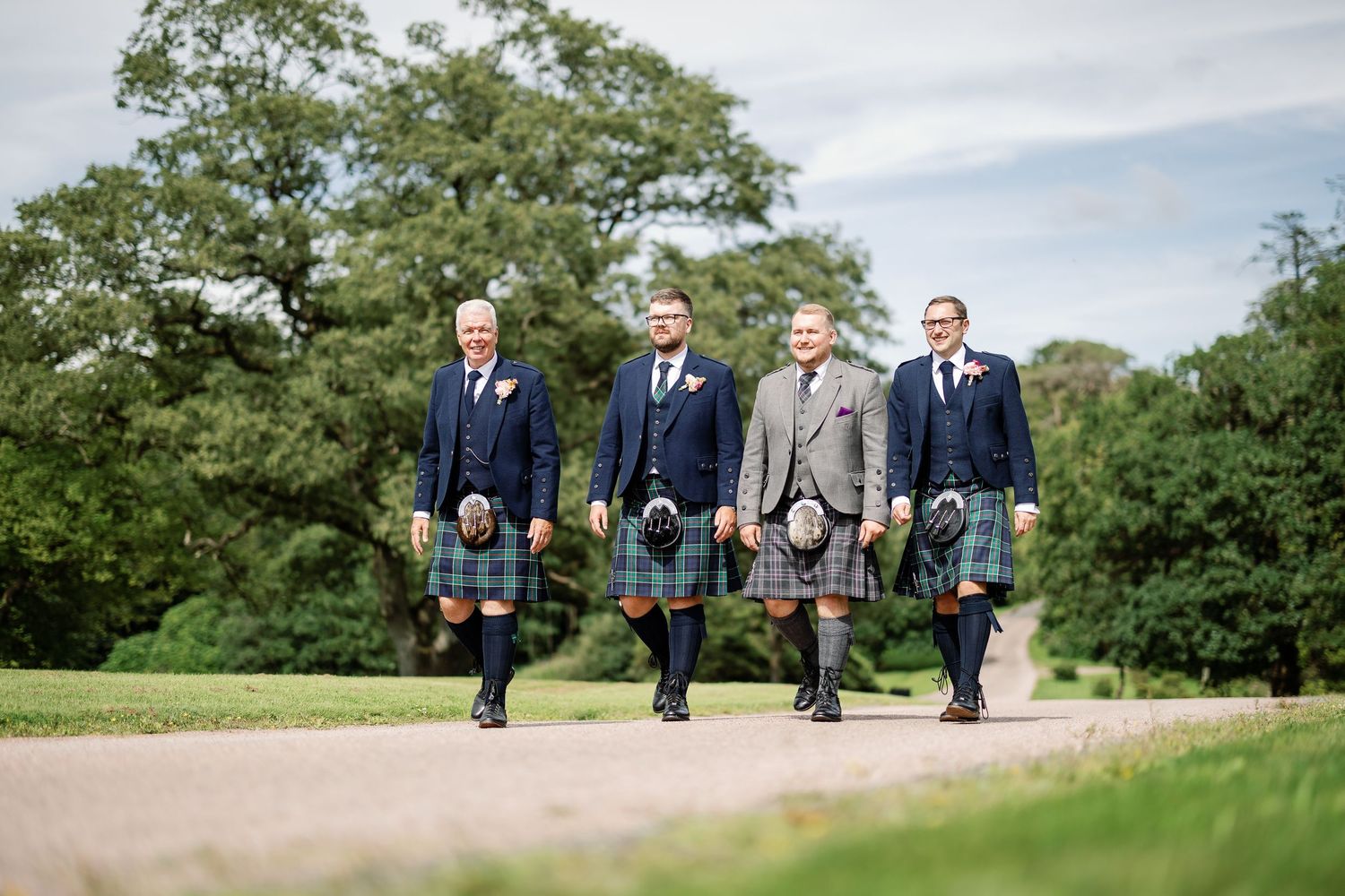Groomsmen in traditional Scottish kilts and suits walk together on a path surrounded by green trees on a cloudy day.