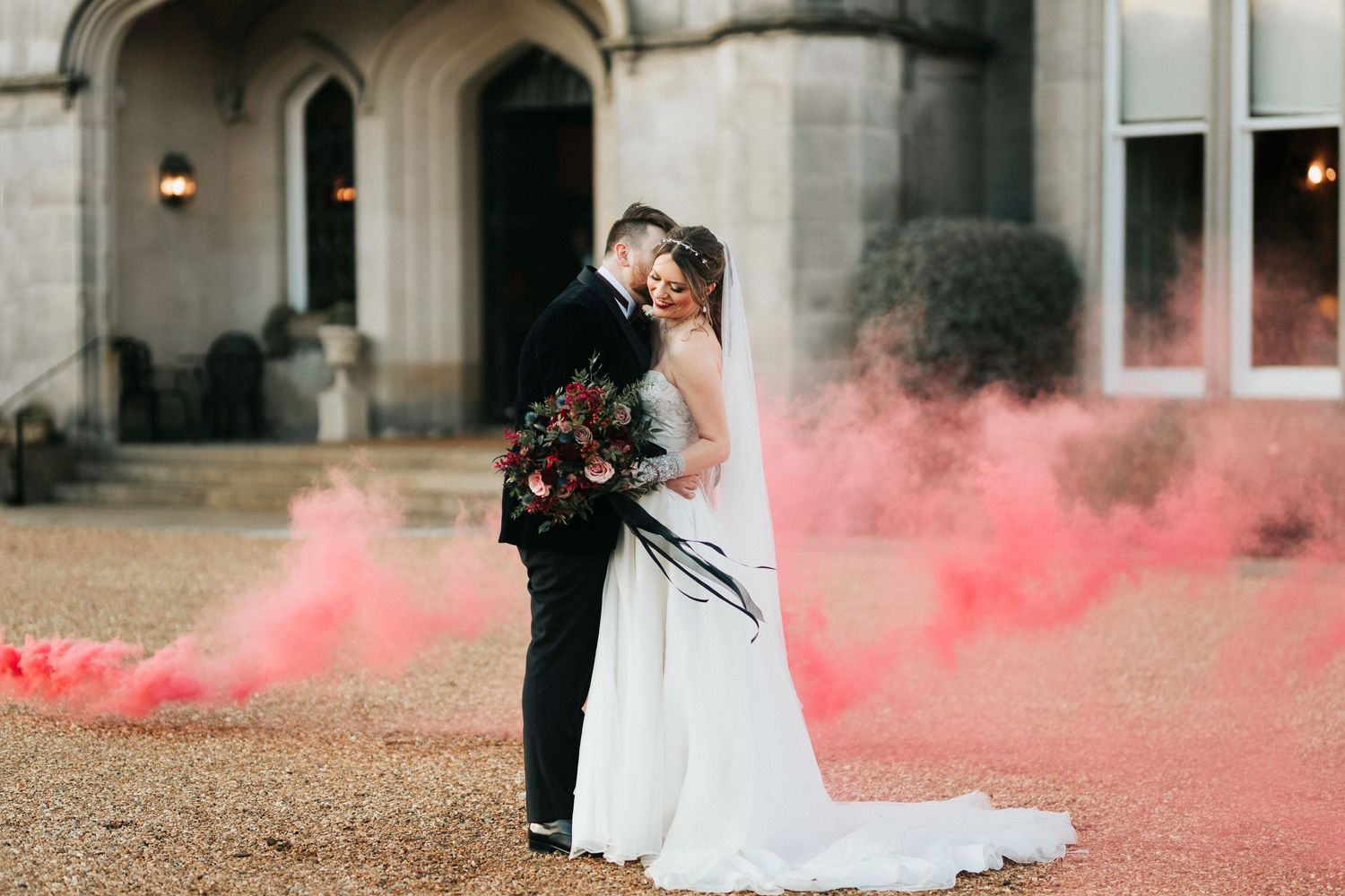 A romantic wedding couple embraces on a gravel driveway with red smoke bombs creating a dramatic atmospheric effect.