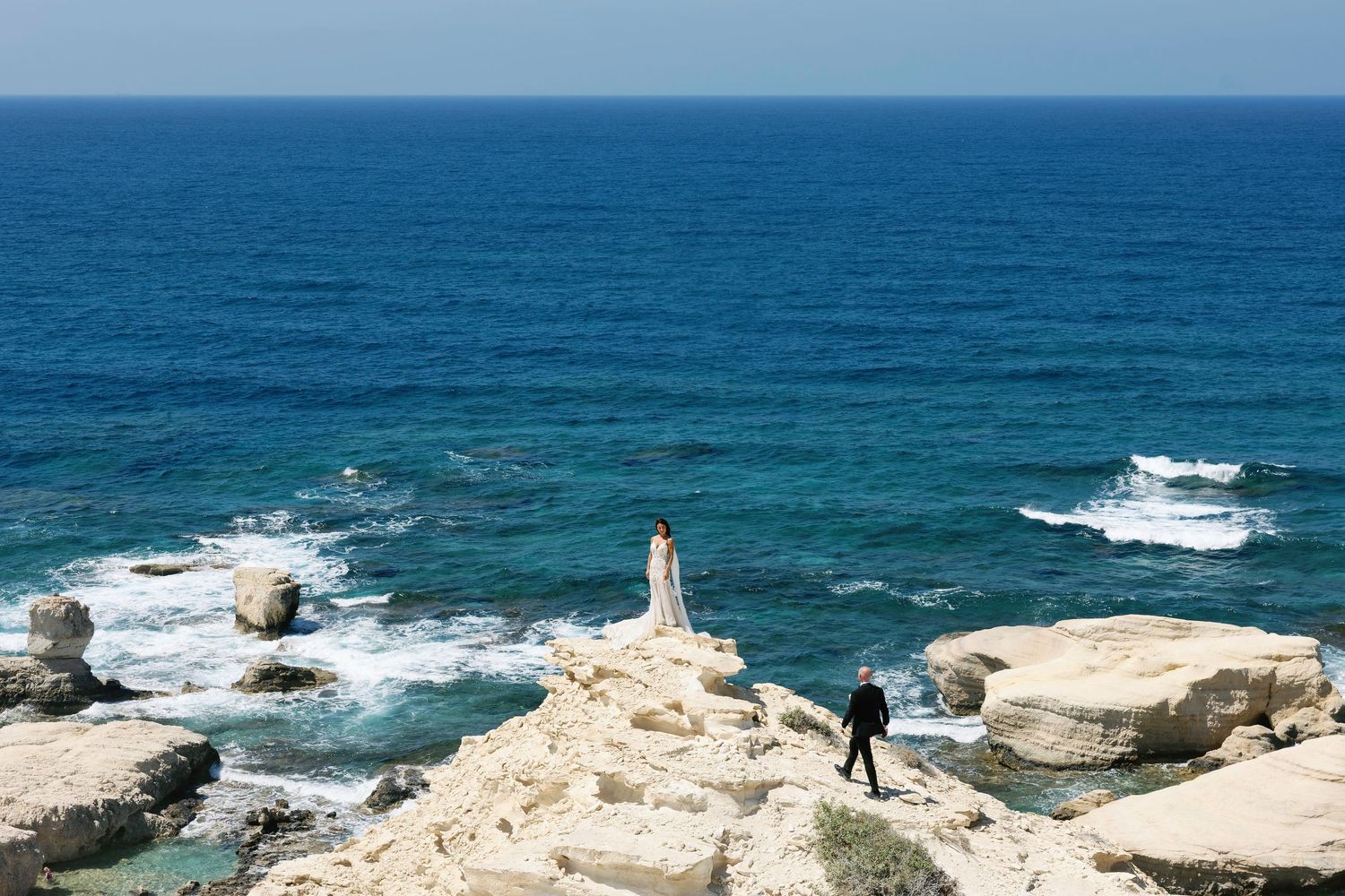 Two people stand on white coastal cliffs overlooking turquoise Mediterranean waters with crashing waves below.