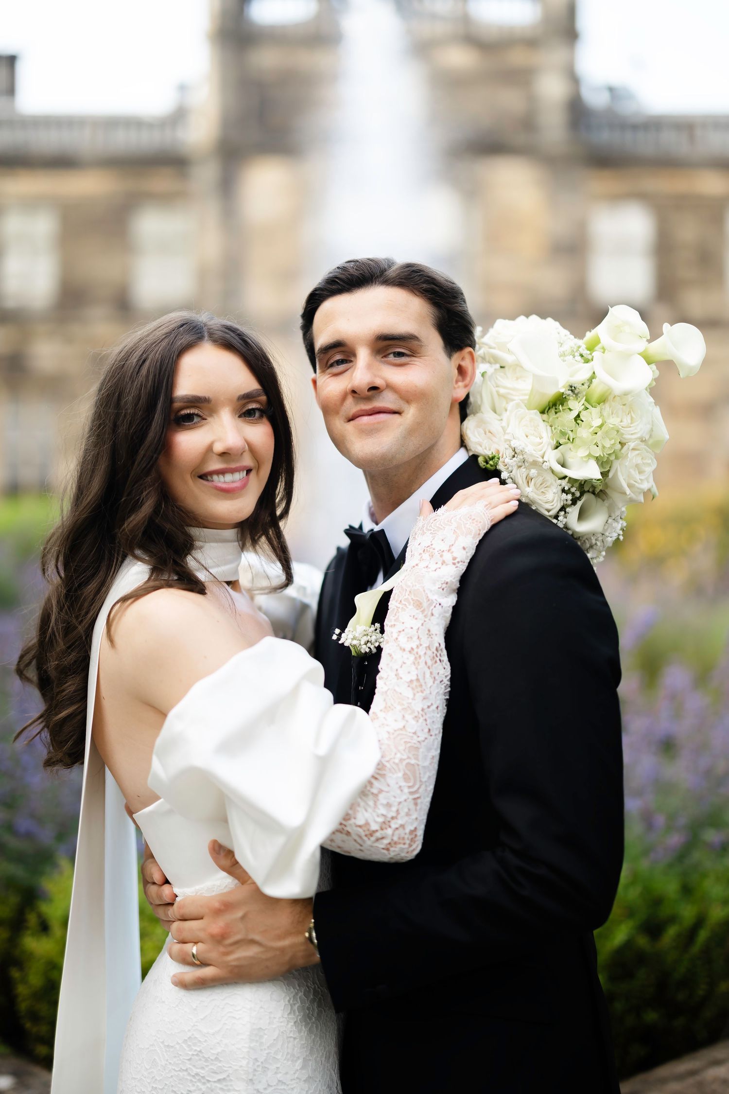 Bride and groom embracing outdoors, she holds white bouquet, wearing off-shoulder gown, he in black tuxedo, fountain backdrop.