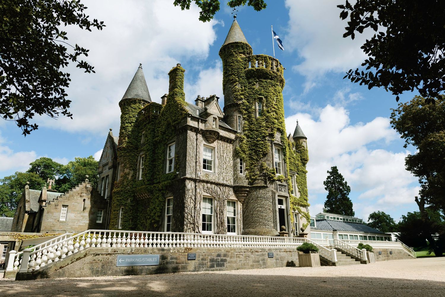 Historic Carlowrie Castle stone castle with ornate turrets and grand staircase stands against blue sky with white clouds framed by trees.