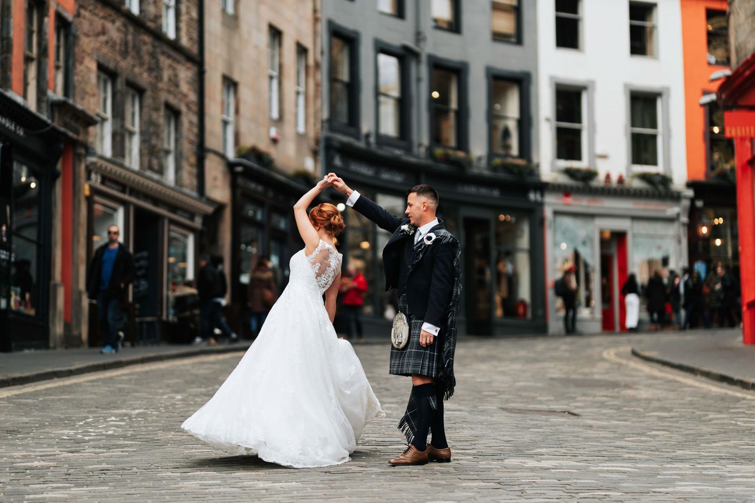 A newlywed couple dances on a historic cobblestone street in Edinburgh, Scotland with traditional Scottish wedding attire.