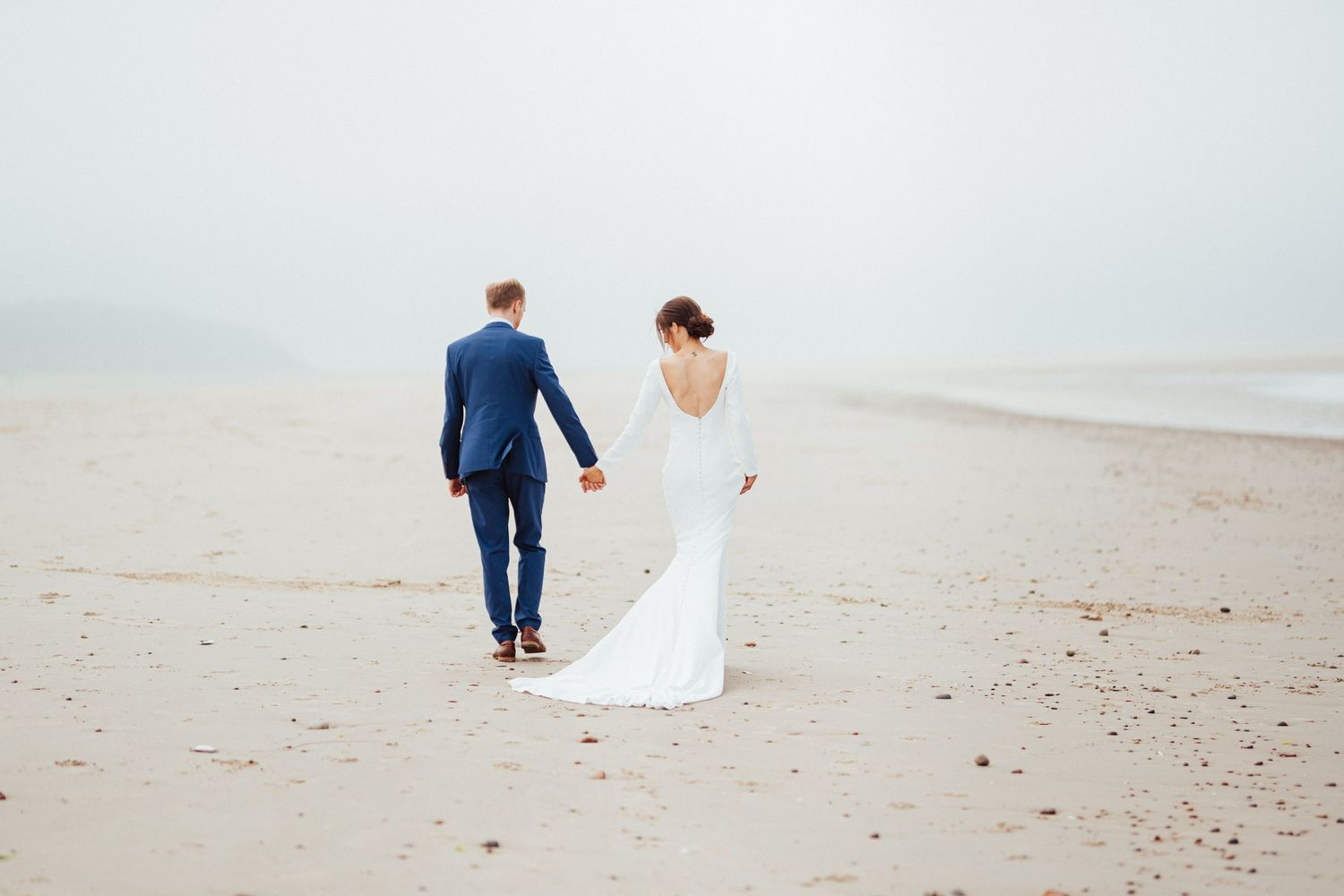 A couple in wedding attire walks together along a misty beach shoreline.