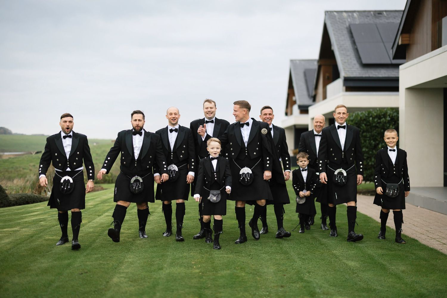 Groomsmen in black tuxedos and kilts pose together on a lush green lawn for formal wedding photos.