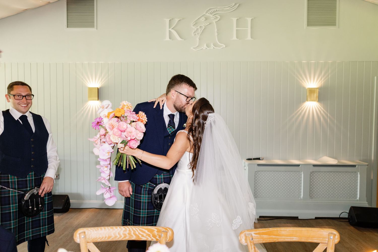 A romantic wedding moment at a venue with a white wall displaying 'K H' lettering and decorative wall sconces.