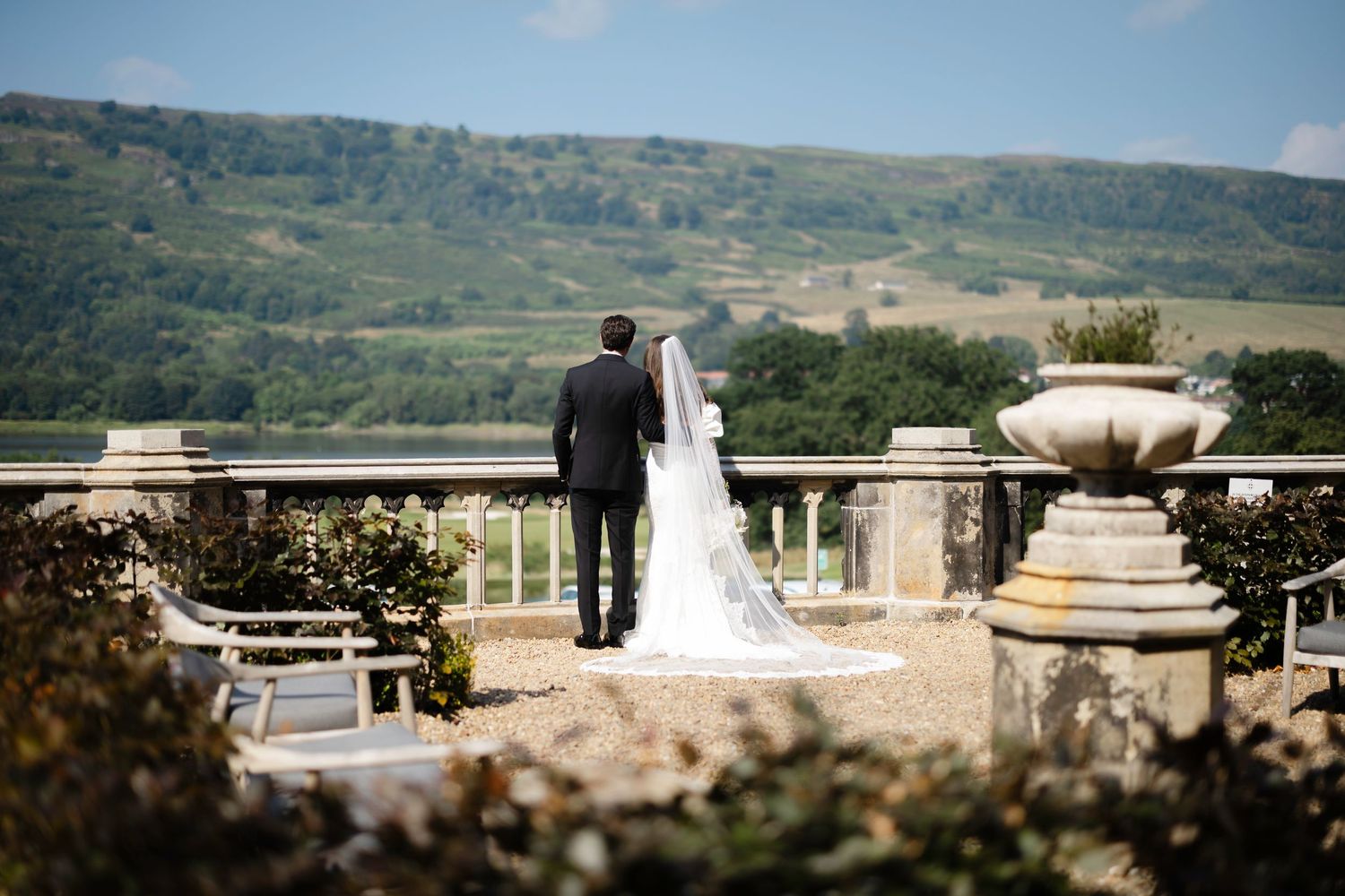 Bride and groom stand on elegant stone terrace overlooking lush green hills on their wedding day.