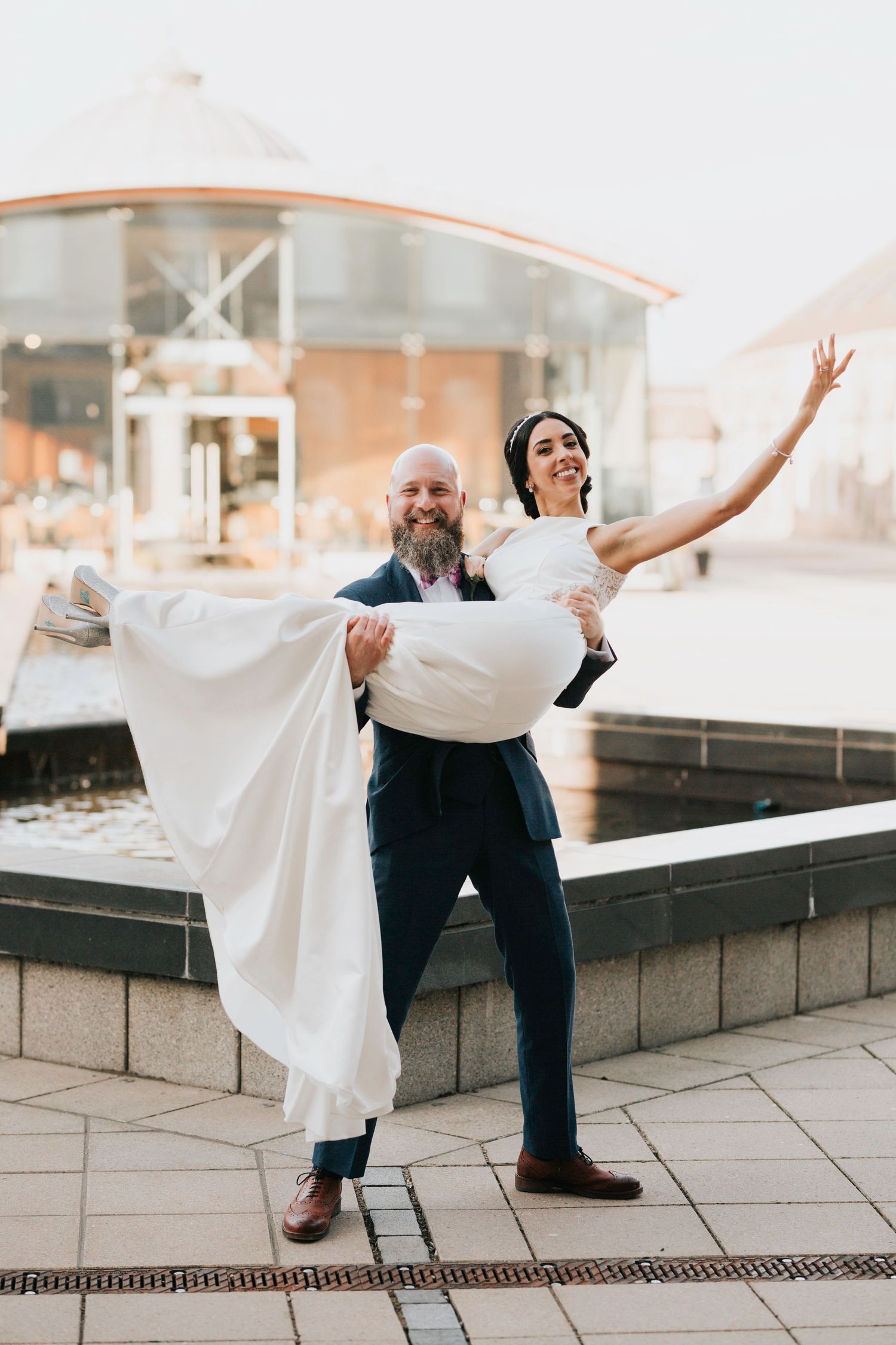 A groom carries his bride in a white wedding dress as she joyfully extends her arm outside an urban building at sunset.