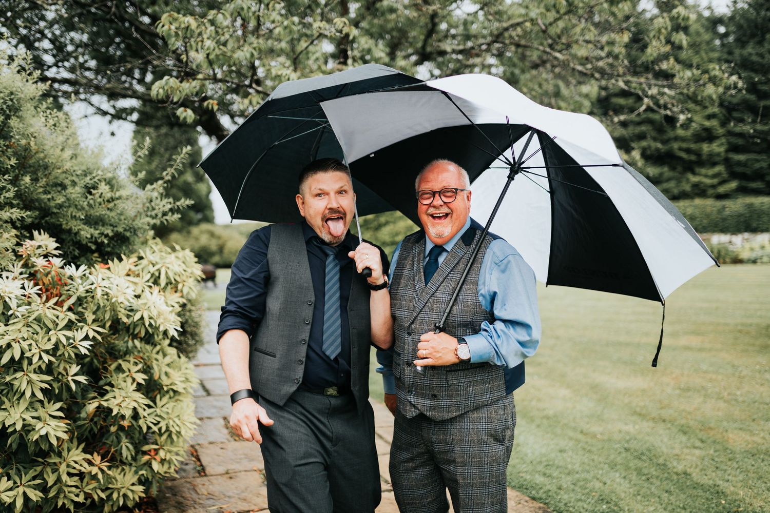 Two people in formal attire sharing a laugh while standing under black and white umbrellas on a green lawn.