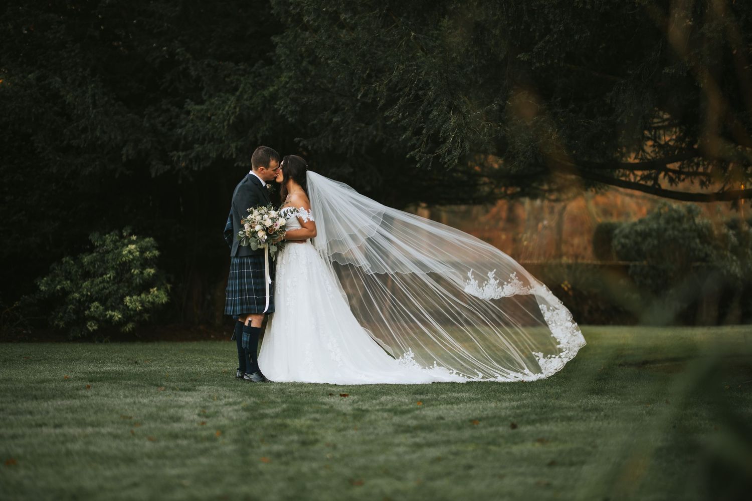 A romantic wedding couple embraces outdoors while the bride's long veil flows dramatically in the wind.