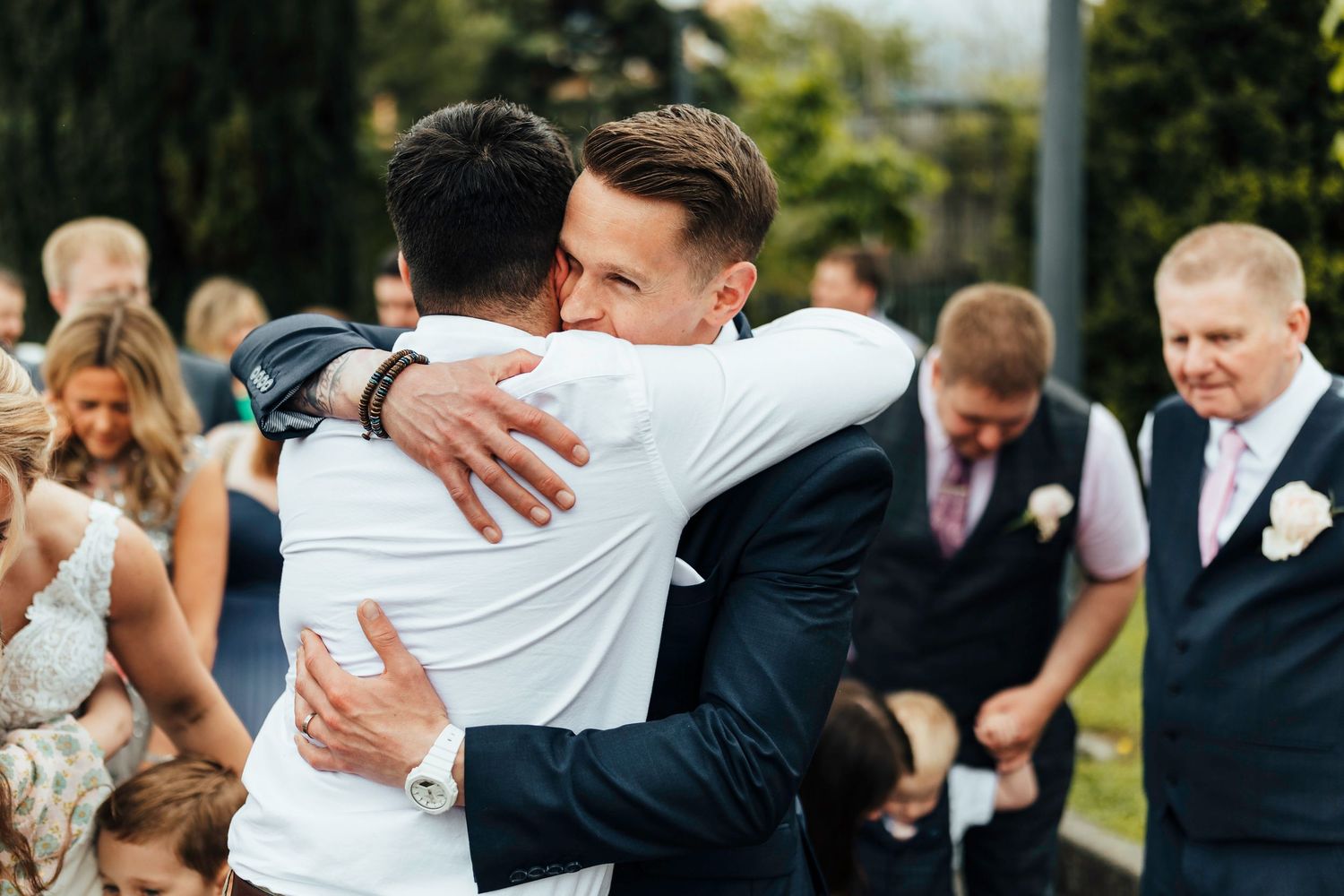 Two people embrace warmly at an outdoor wedding ceremony while guests look on in the background.