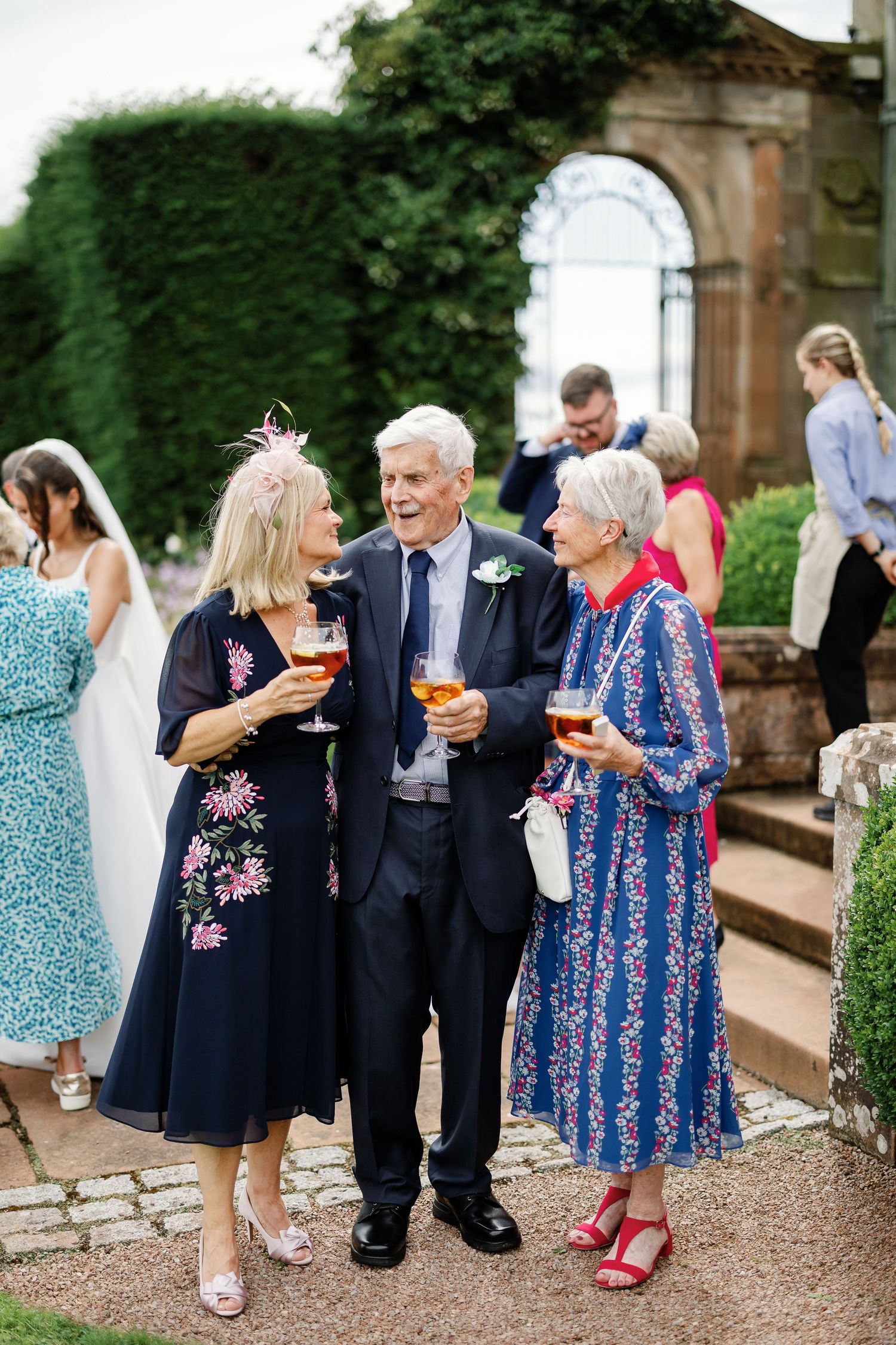 Wedding guests in formal attire chat and mingle during an outdoor garden reception.