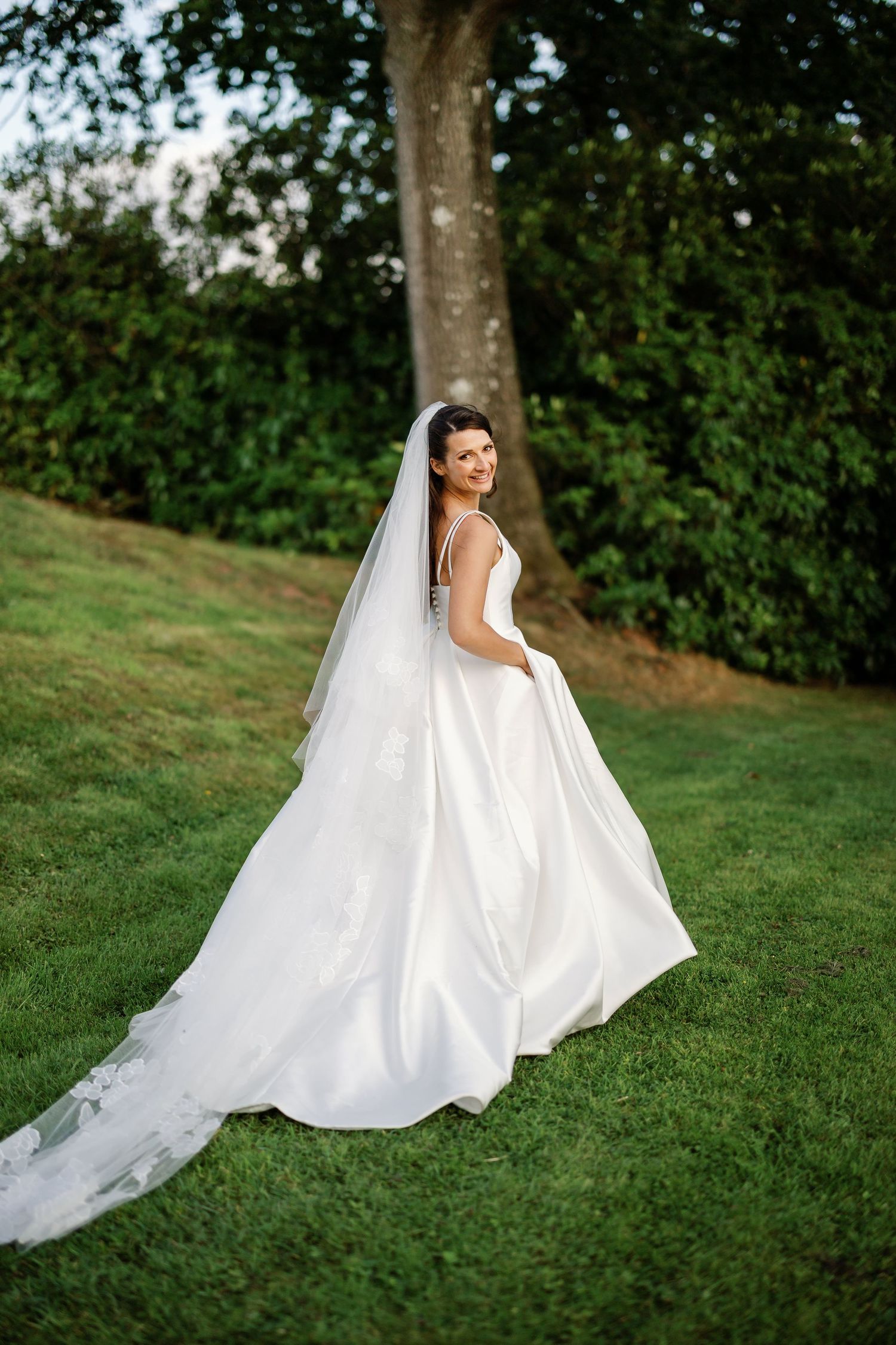 A bride in a flowing white wedding gown poses gracefully on a green grassy hill during an outdoor photo session.