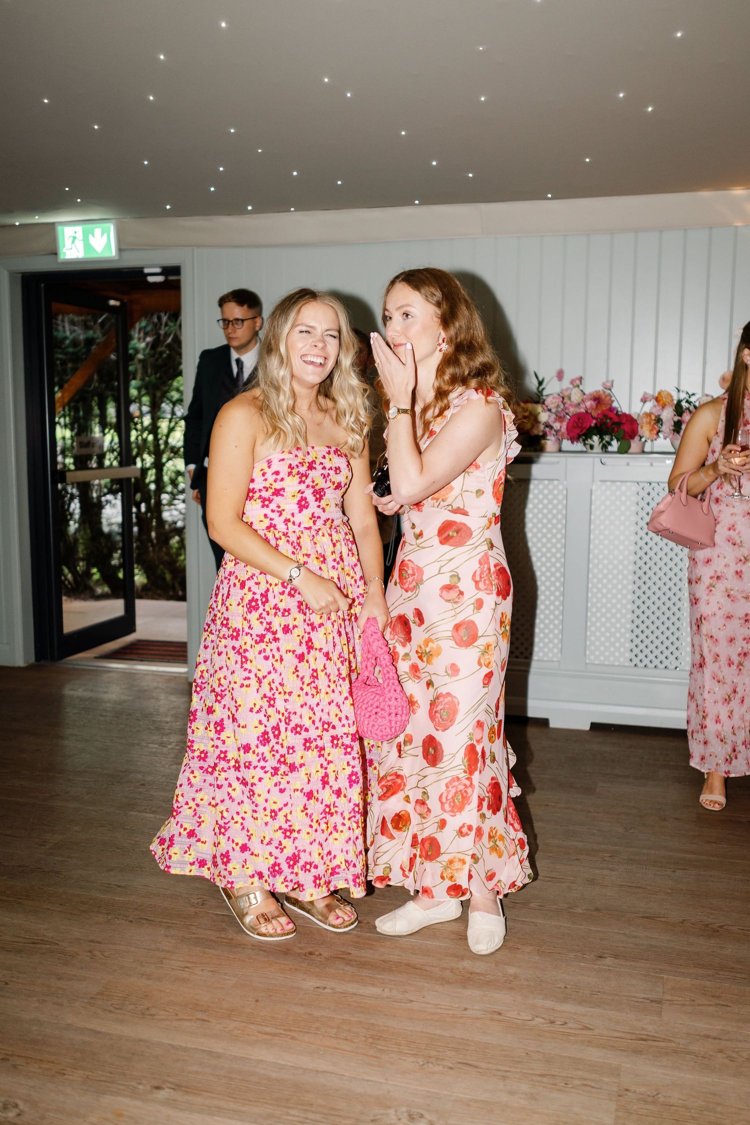 Two women in floral summer dresses share a laugh at an indoor event with twinkling lights overhead.