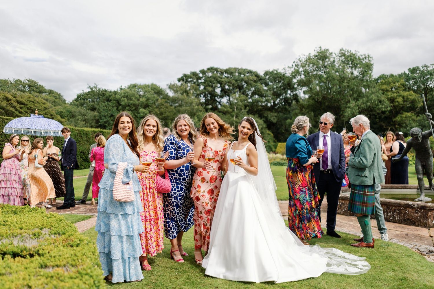 Wedding party members in colorful dresses and suits gather outdoors for group photos at a garden celebration.