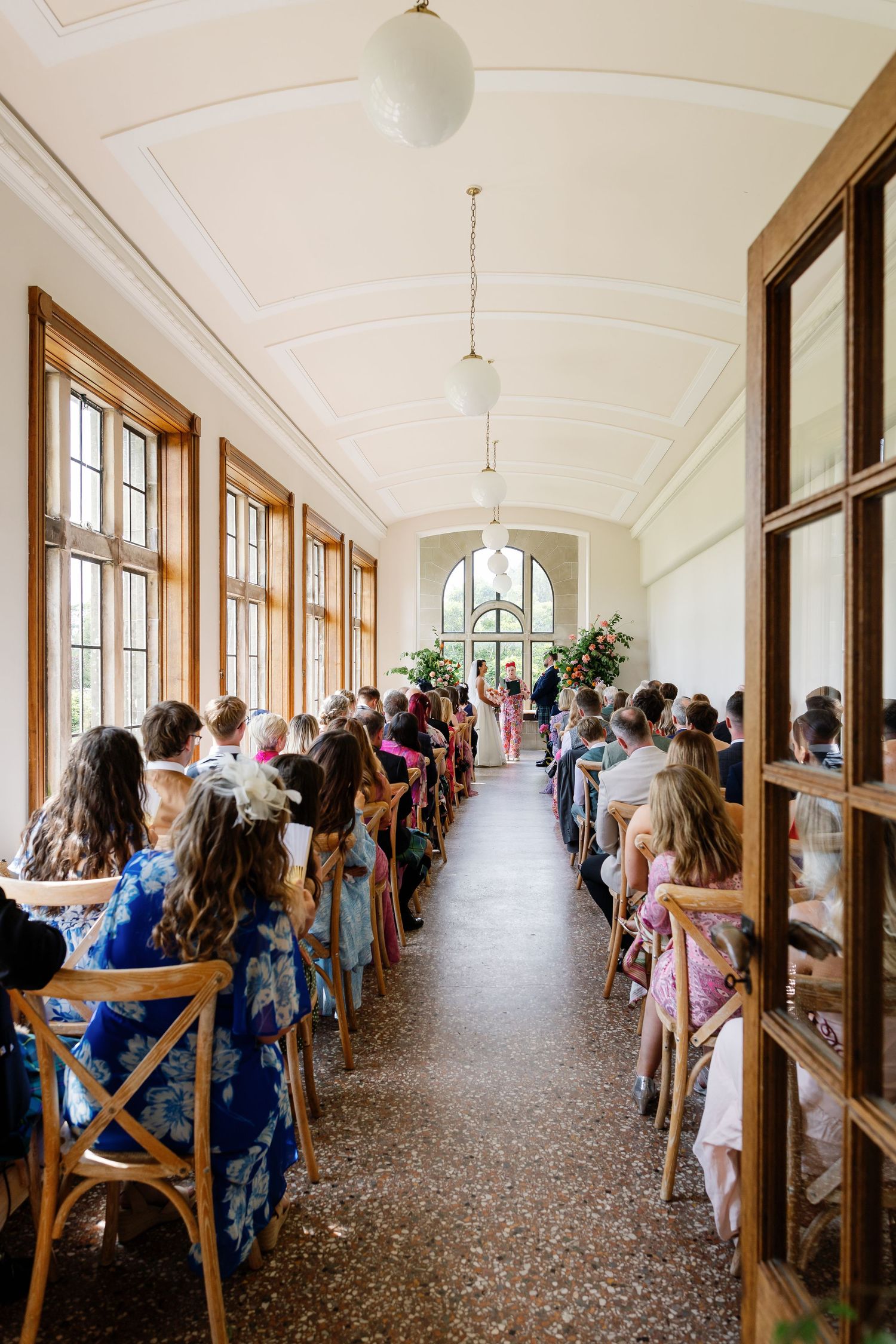 A long corridor with guests seated on benches along windows during an indoor ceremony.
