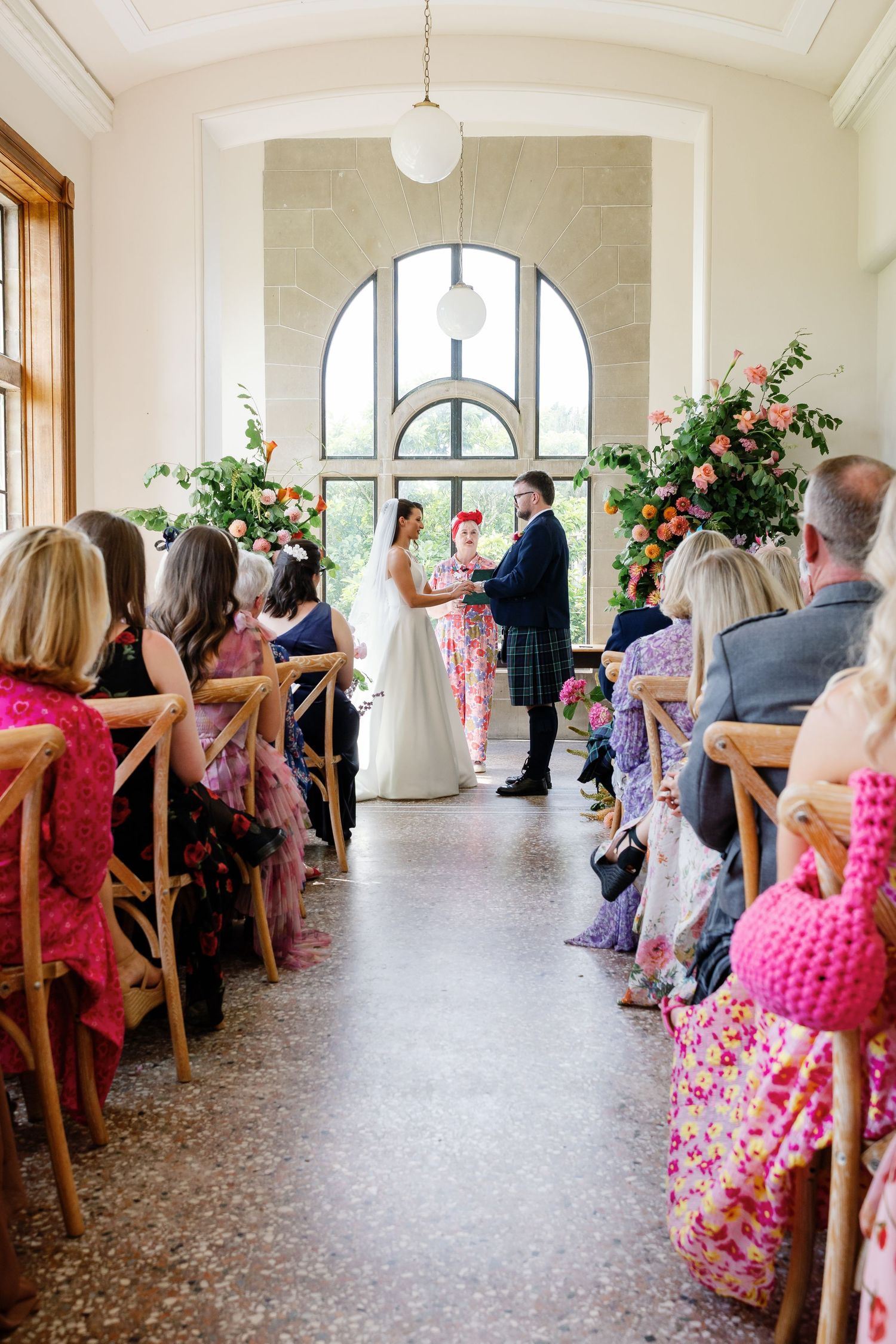 Wedding guests seated in wooden chairs along a bright hallway with arched windows and floral decorations watch a ceremony.
