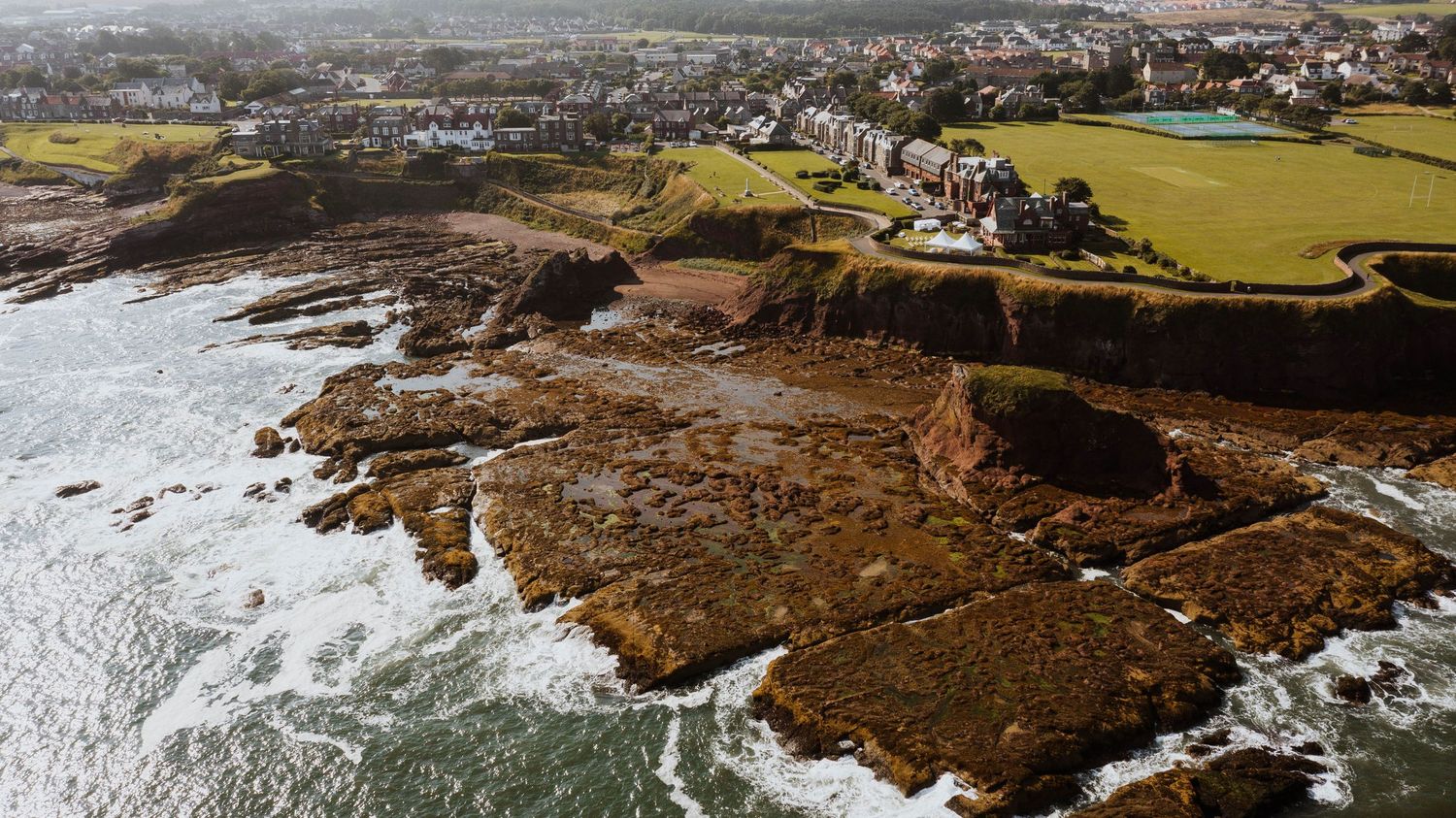 Aerial view of rugged coastal cliffs with houses perched along the rocky shoreline and green fields stretching inland.