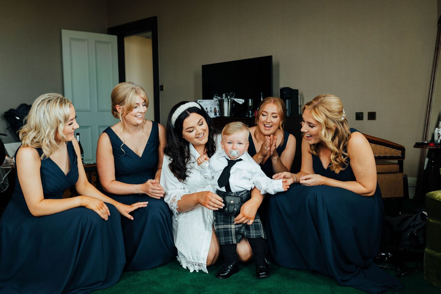 Wedding party in navy blue dresses gathers on floor sharing joyful moment with small child in formal attire.