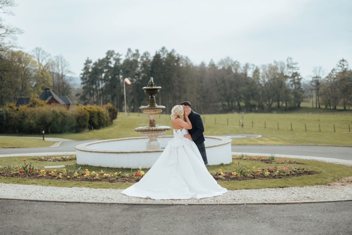 A couple shares a romantic moment in their wedding attire near a landscaped garden roundabout at a countryside venue.