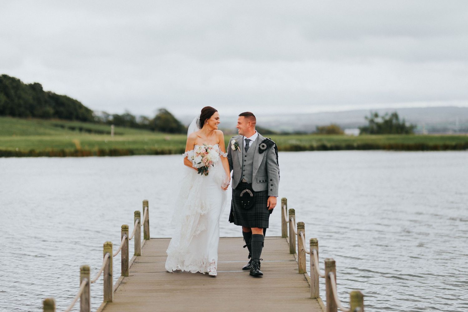 A bride and groom share a romantic moment on a wooden dock overlooking a serene lake and countryside.