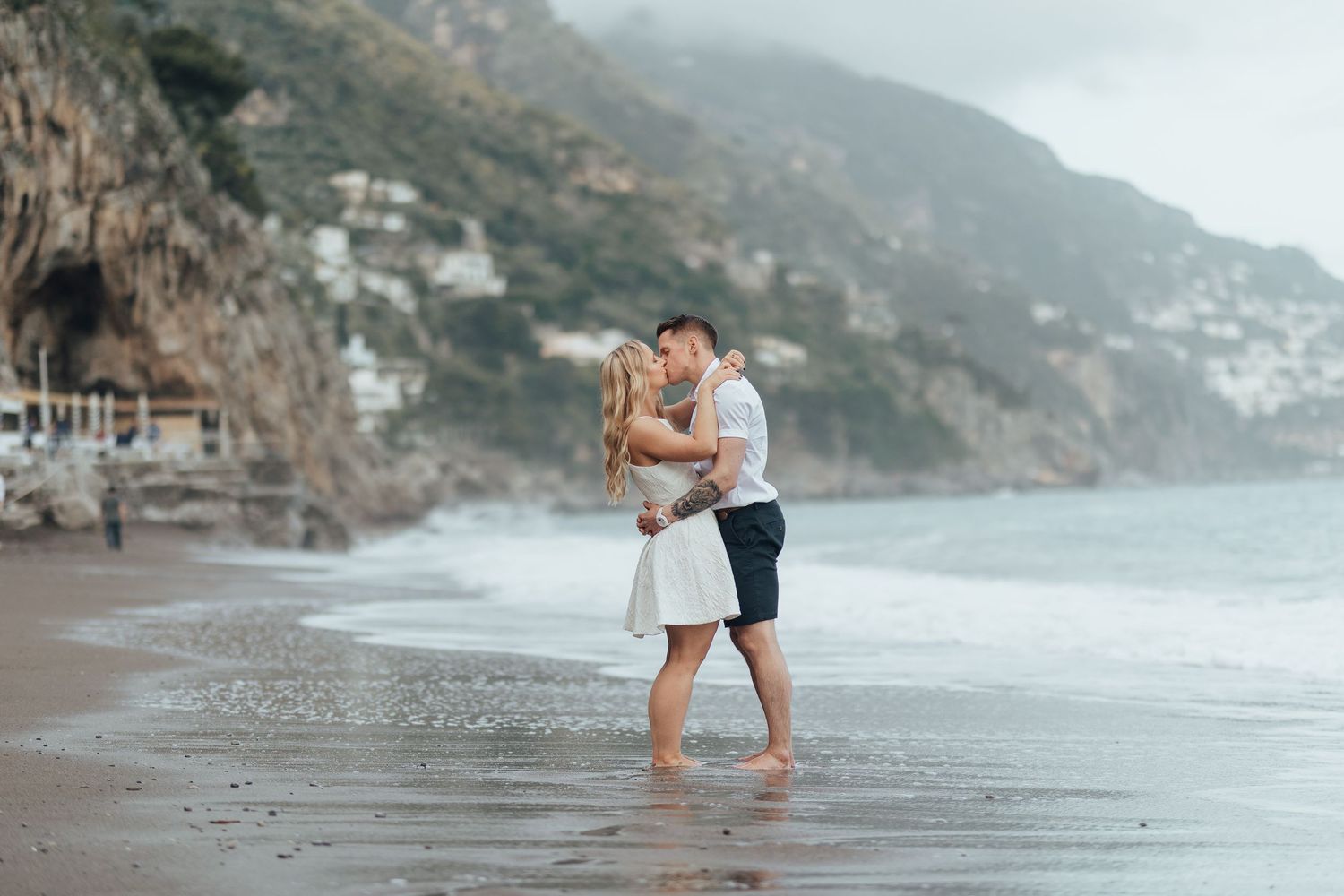 Two romantic figures embrace while standing barefoot on a misty beach with dramatic coastal cliffs in the background.